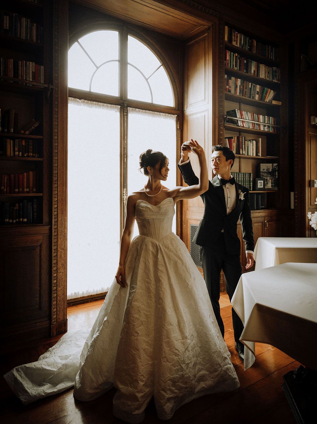 Elegant bride and groom dancing in a grand library with arched windows and bookshelves.