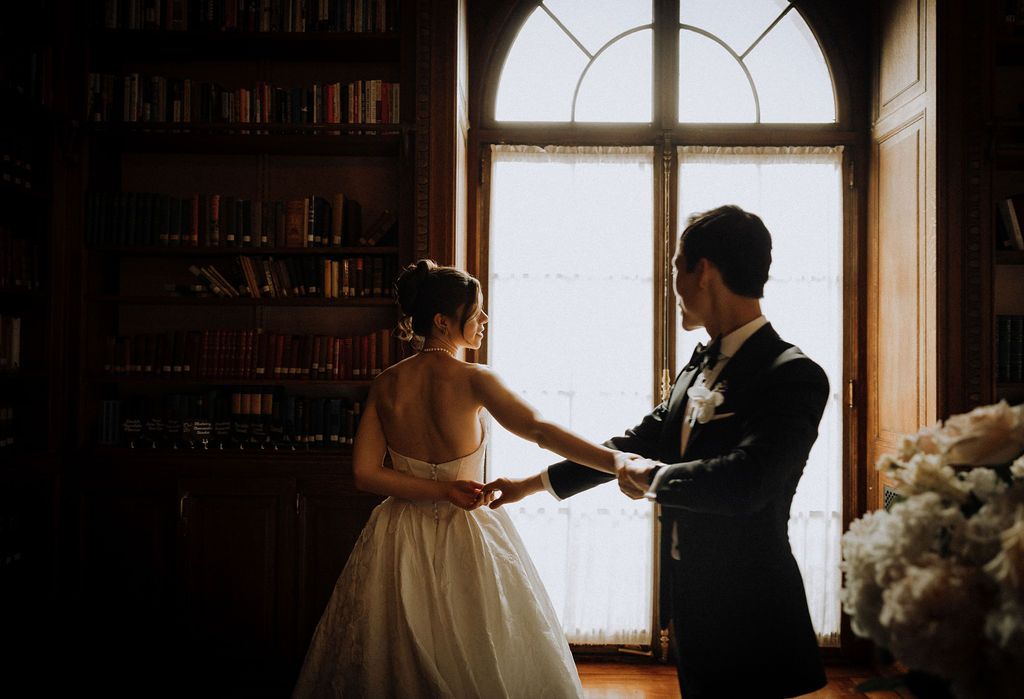 Bride and groom holding hands in a dimly lit library with arched windows and bookshelves during wedding ceremony.