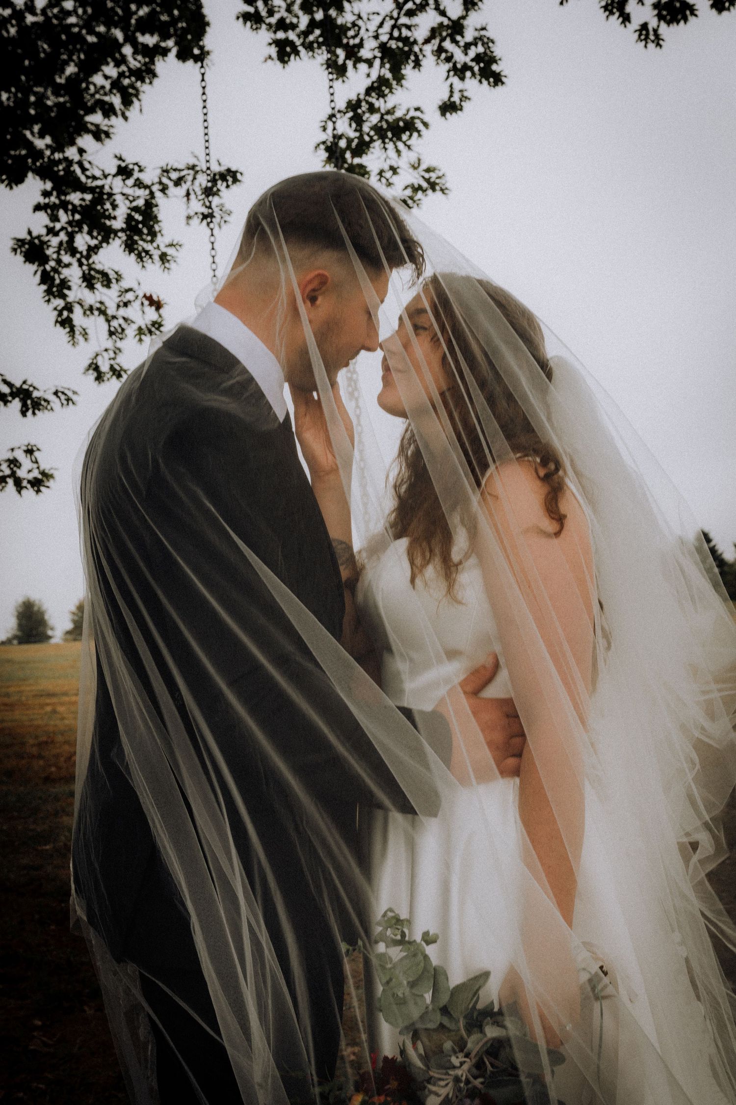An intimate wedding portrait showing a tender moment under a delicate veil in a dreamy outdoor setting.