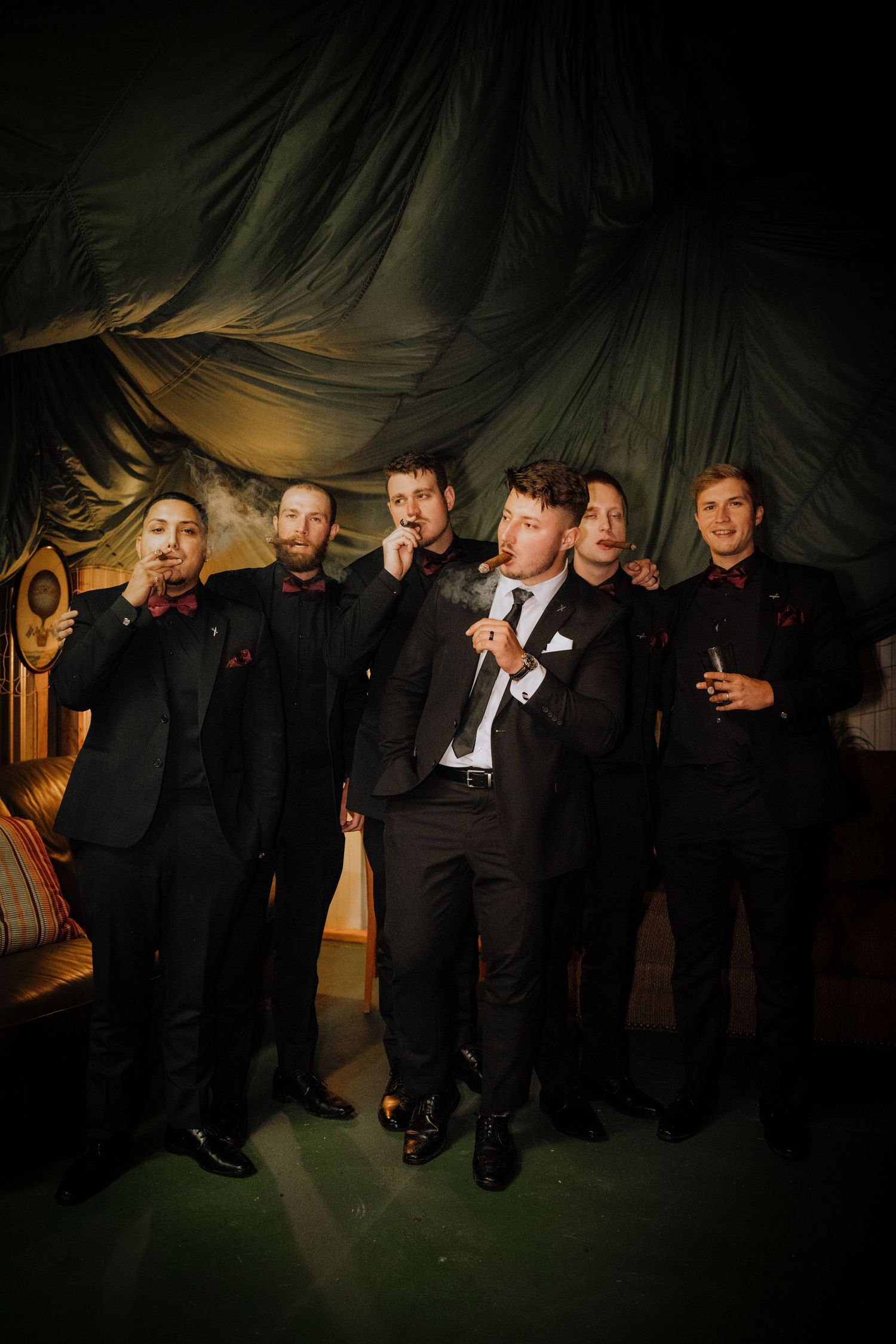 Wedding party in black suits posed against dark backdrop with dramatic lighting.