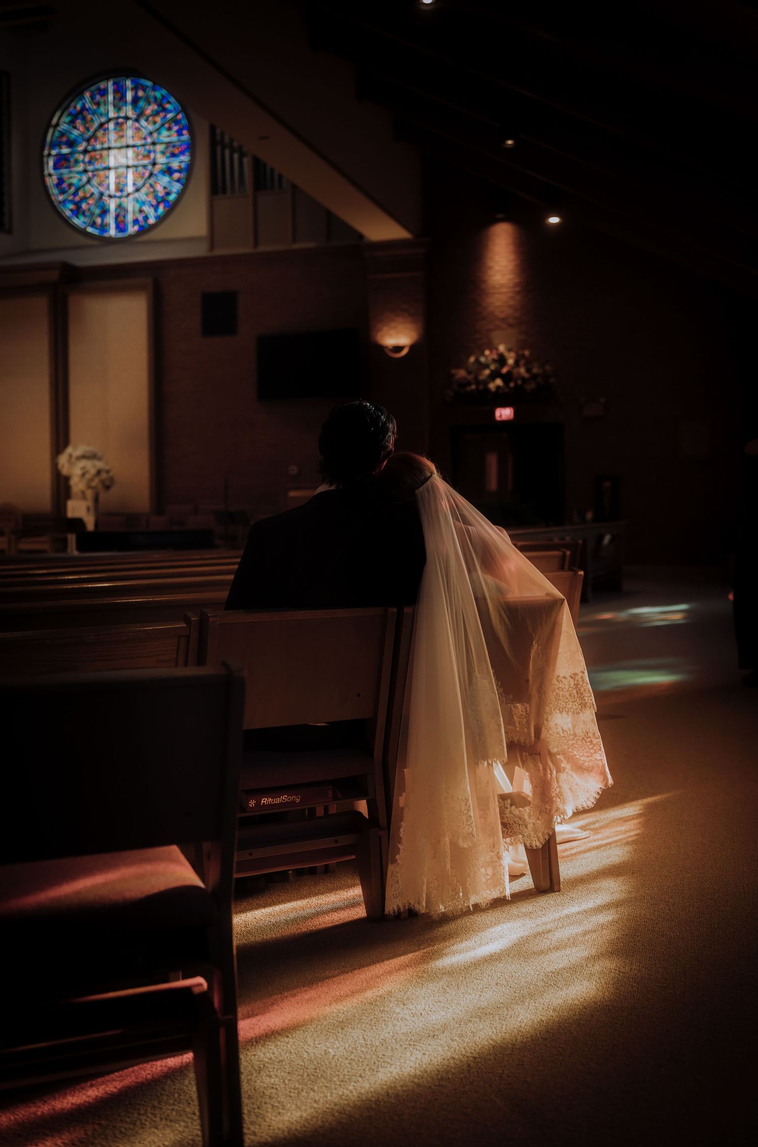 Sunlight streams through stained glass windows onto a wedding dress in a dimly lit church.