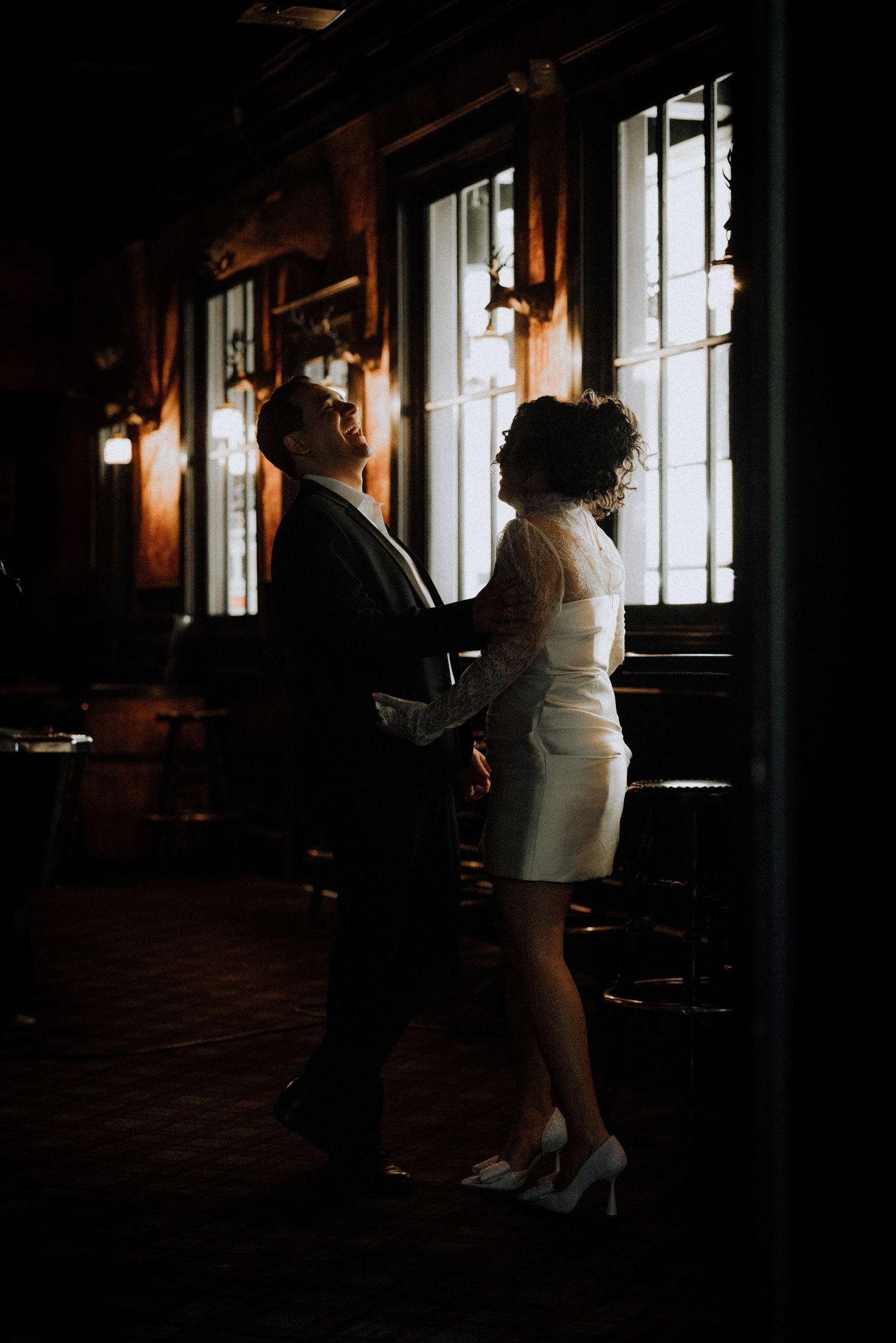 editorial-engagement-photograBride and groom share a romantic moment in a dimly lit vintage venue, silhouetted against warm glowing windows.
