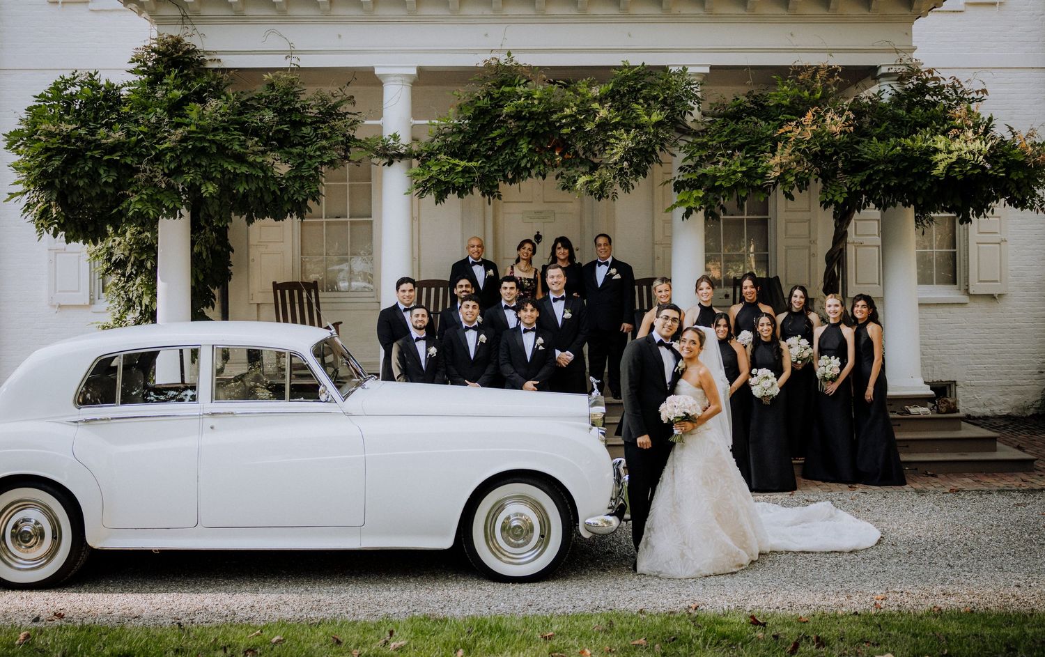 Wedding party poses with classic white Rolls-Royce before elegant white manor, bride and groom centered among guests.