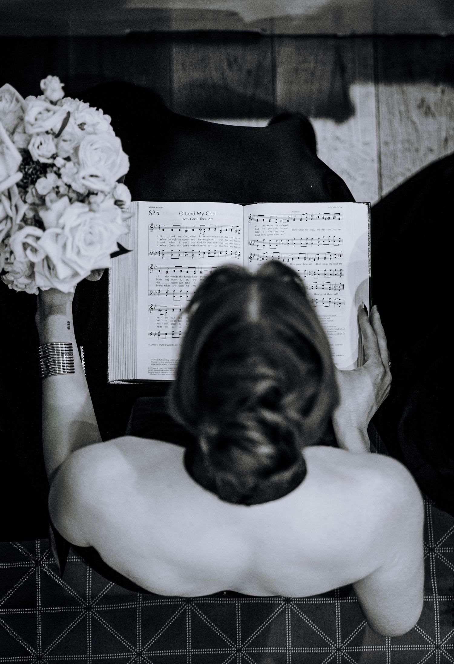 Black and white photo of bride holding bouquet reading sheet music book, viewed from above.