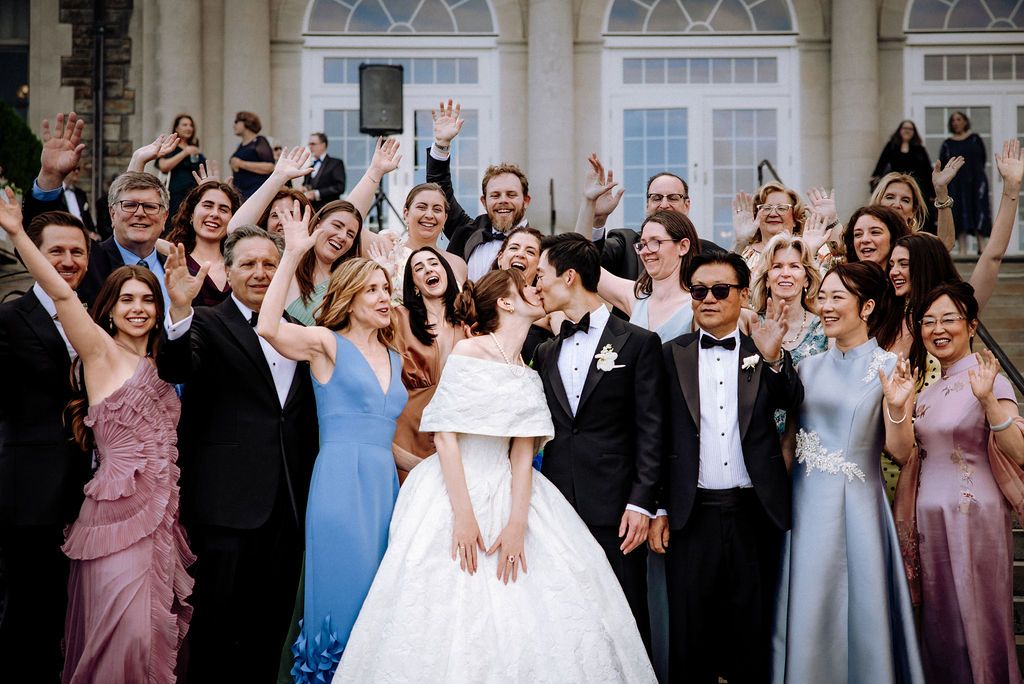 Bride and groom kissing surrounded by cheering wedding guests on steps of elegant venue.
