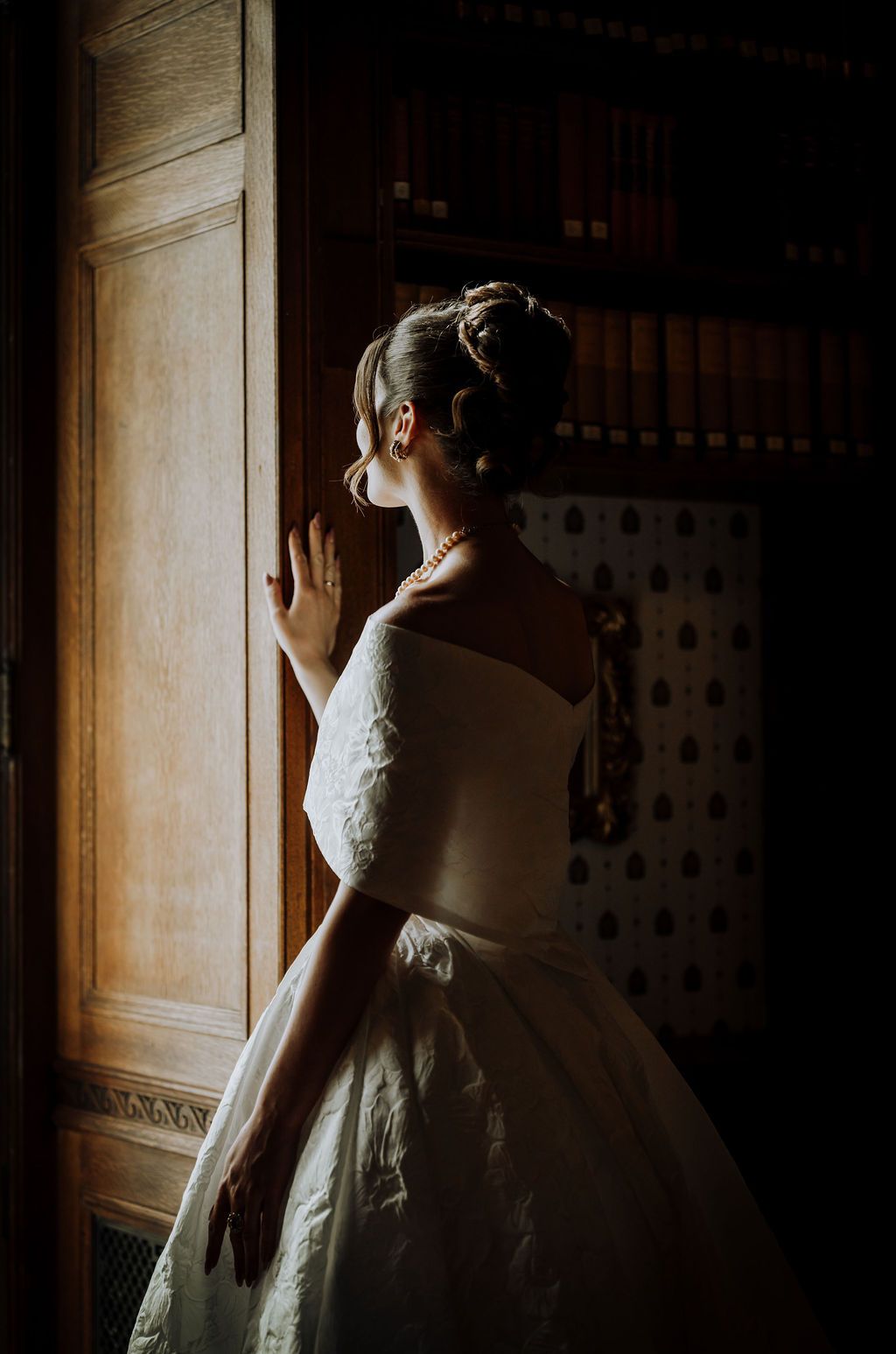 Bride in elegant off-shoulder white gown with updo hairstyle and pearl necklace, gazing out a window in a grand library.