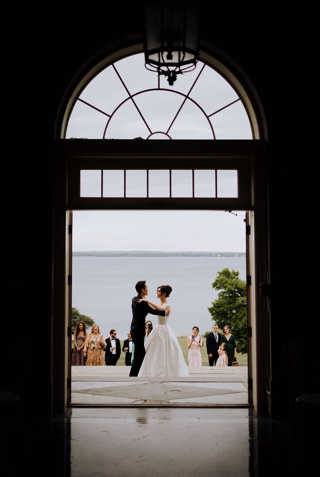 Bride and groom share first dance framed by elegant arched doorway with waterfront view and guests in background.