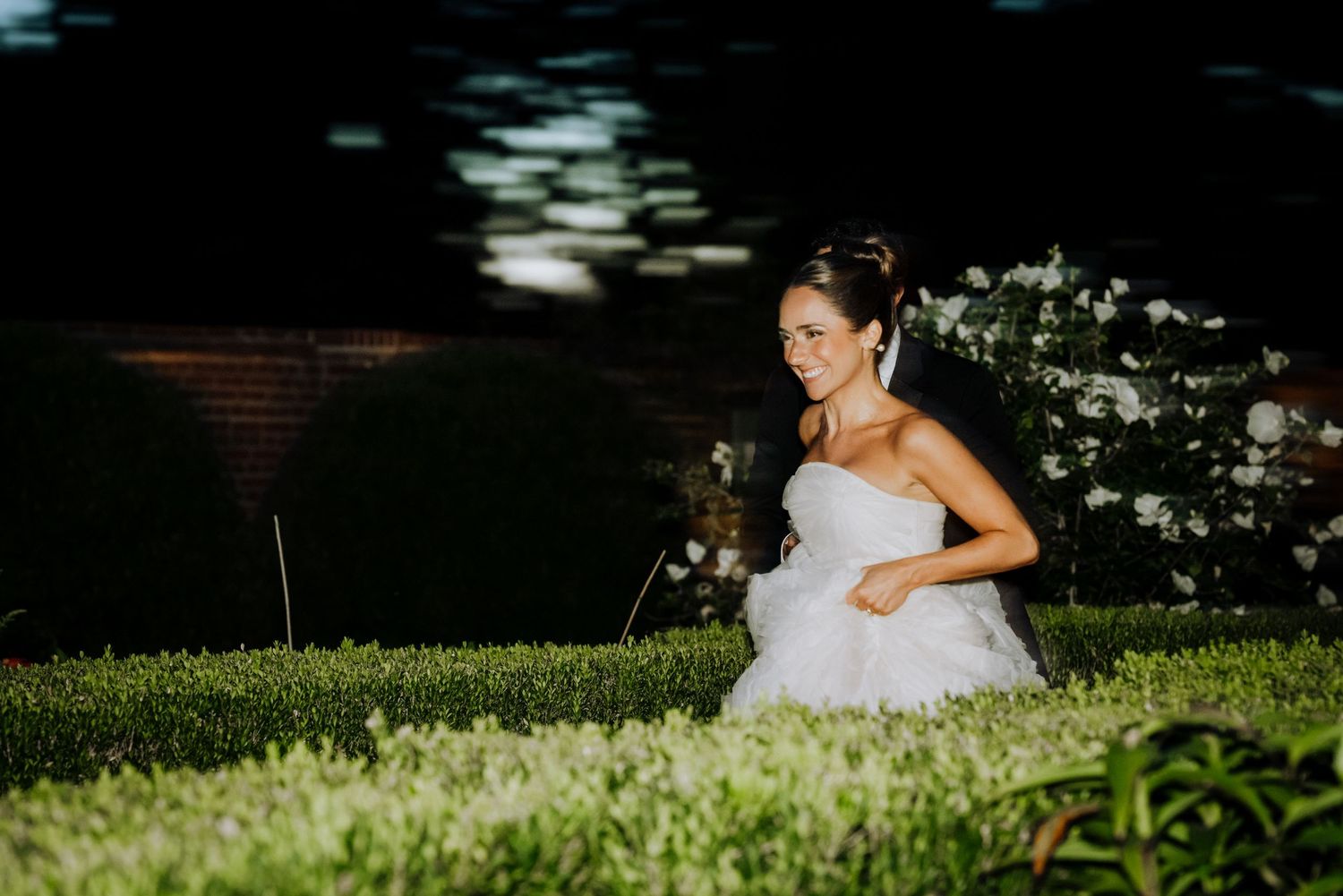 Bride in white wedding gown running through lush green garden at night, smiling joyfully.