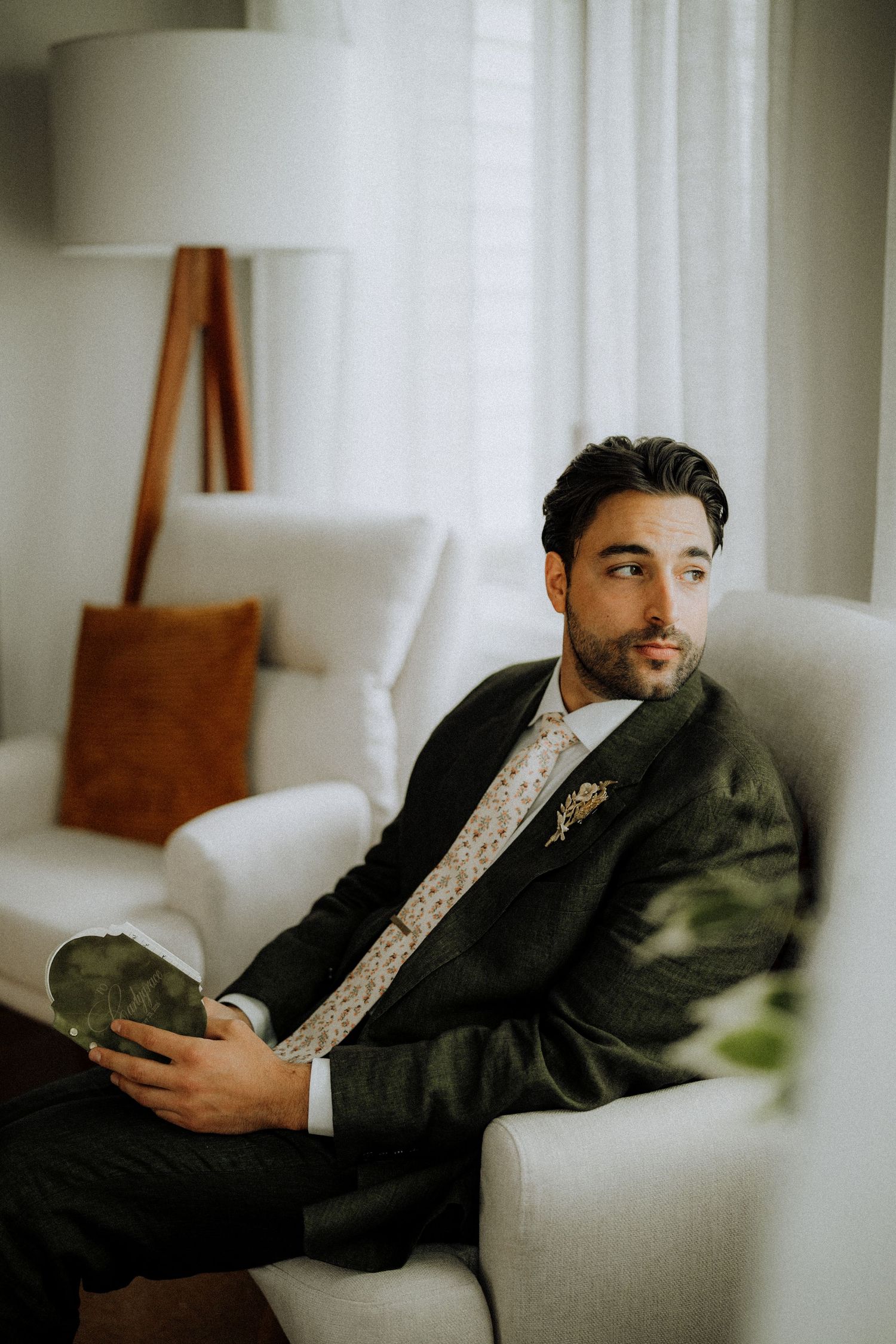 A well-dressed groom in a dark suit and floral tie sits relaxed in a white armchair, gazing thoughtfully toward a window.