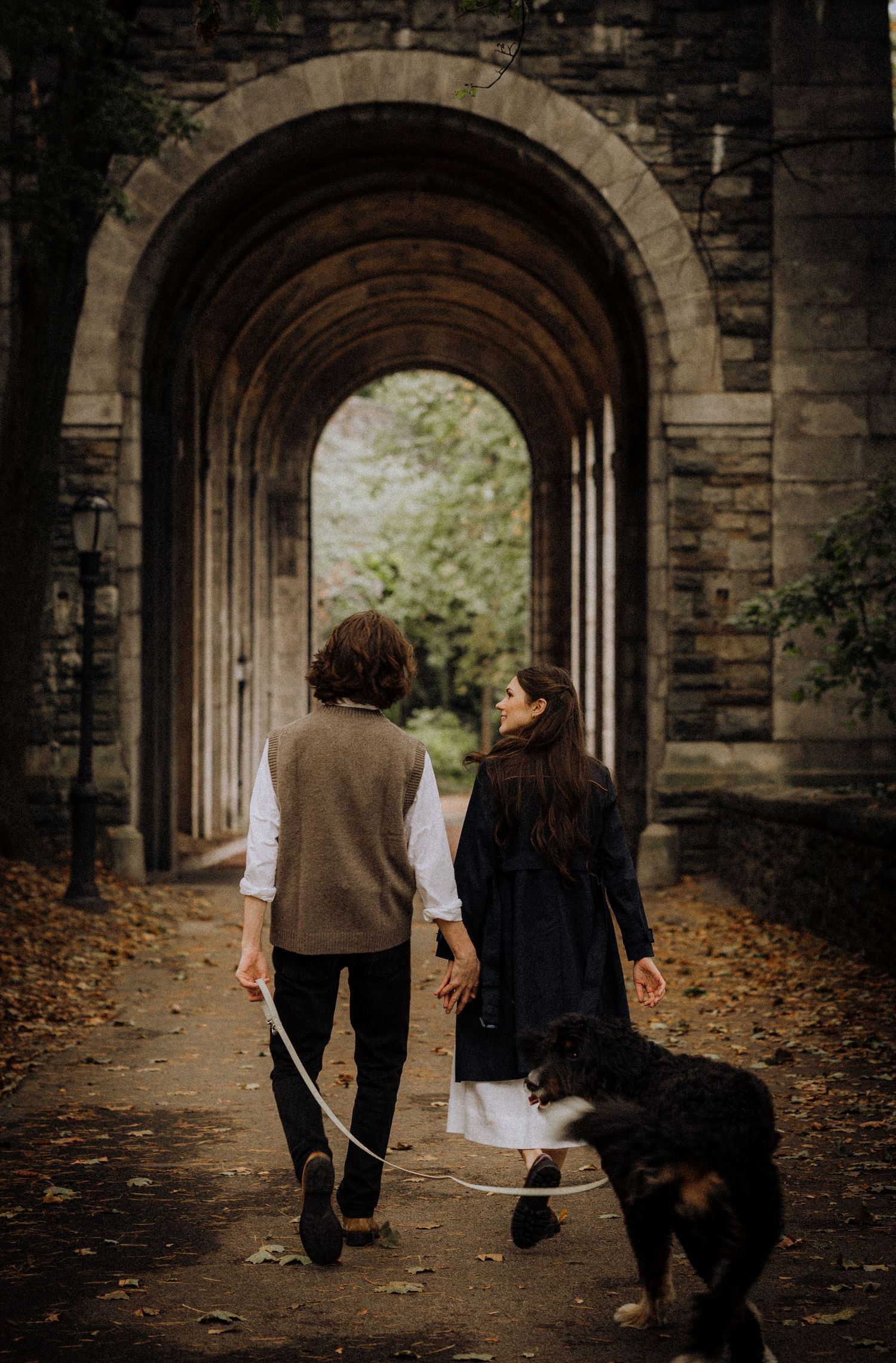 A couple walks with their dog through a beautiful stone archway corridor covered in autumn leaves.