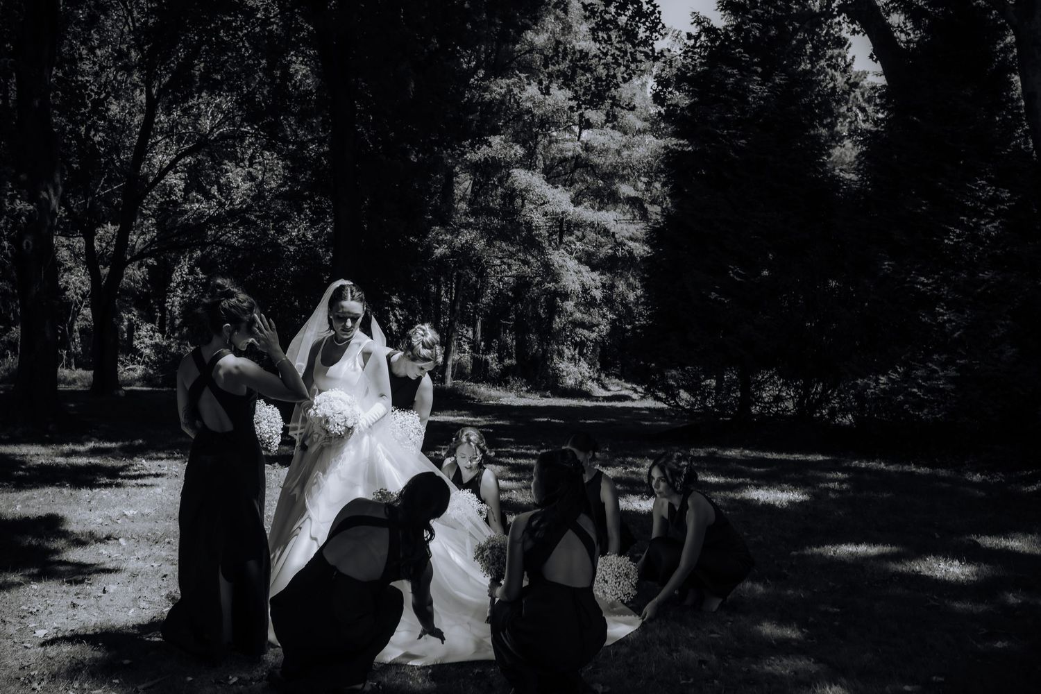 Black and white photo of a bride's dress being arranged by attendants outdoors.
