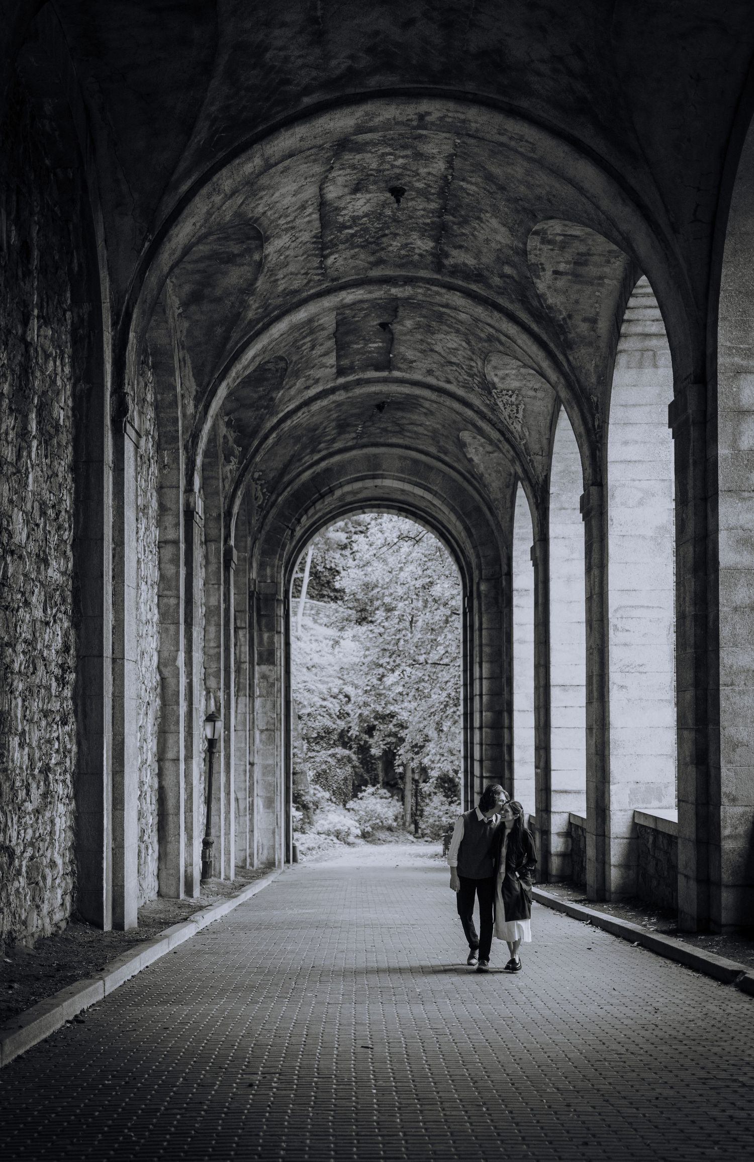 A couple walks through a dramatic stone archway corridor in black and white photography.