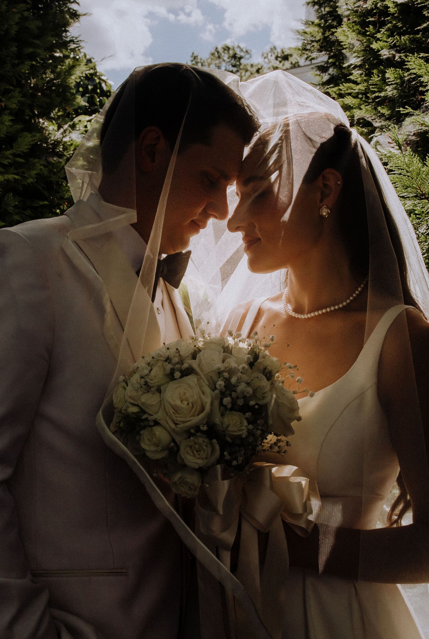 Intimate moment between newlyweds under a veil with white rose bouquet.