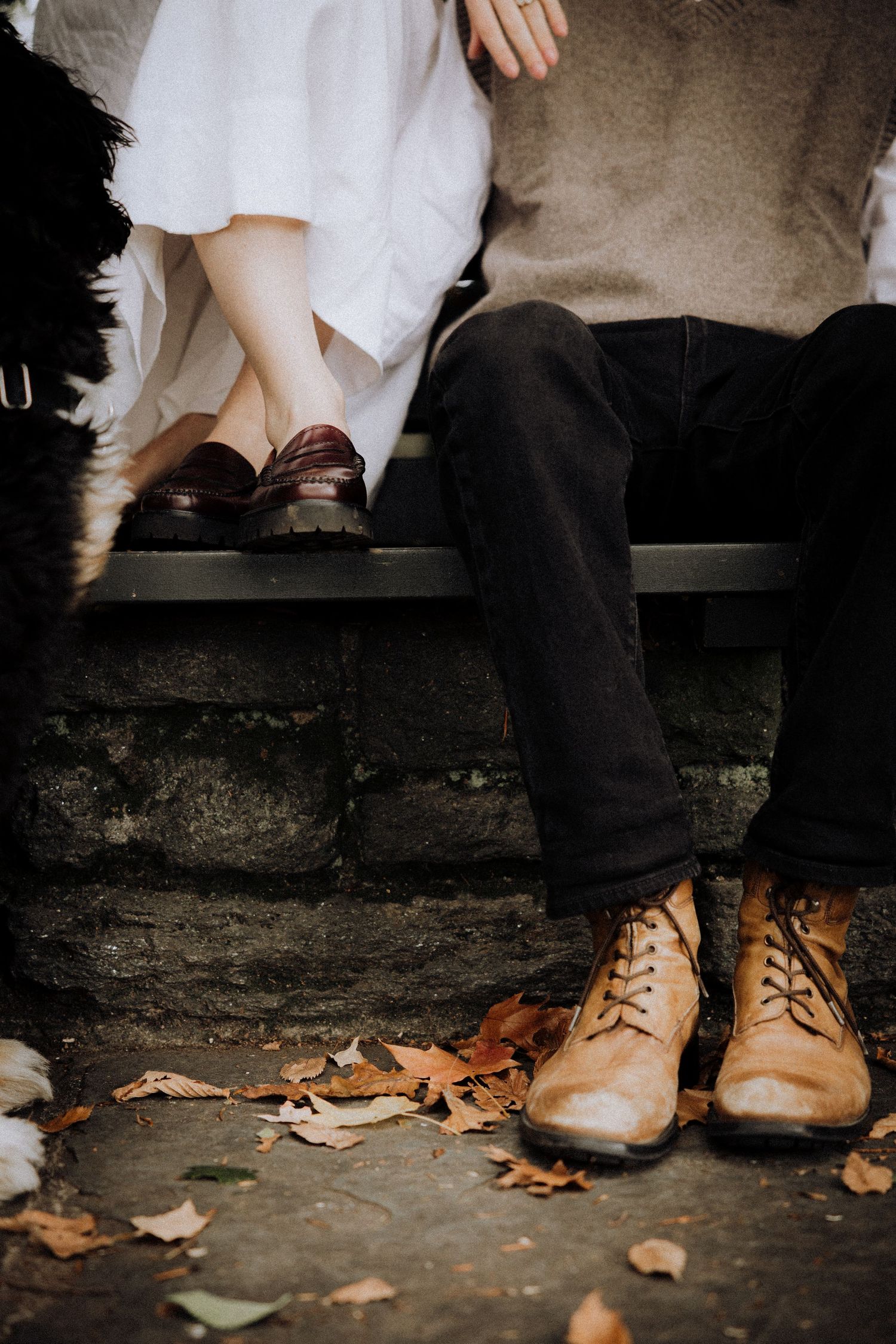 Detail shot of feet wearing brown leather boots sitting on a wooden bench with fallen autumn leaves.