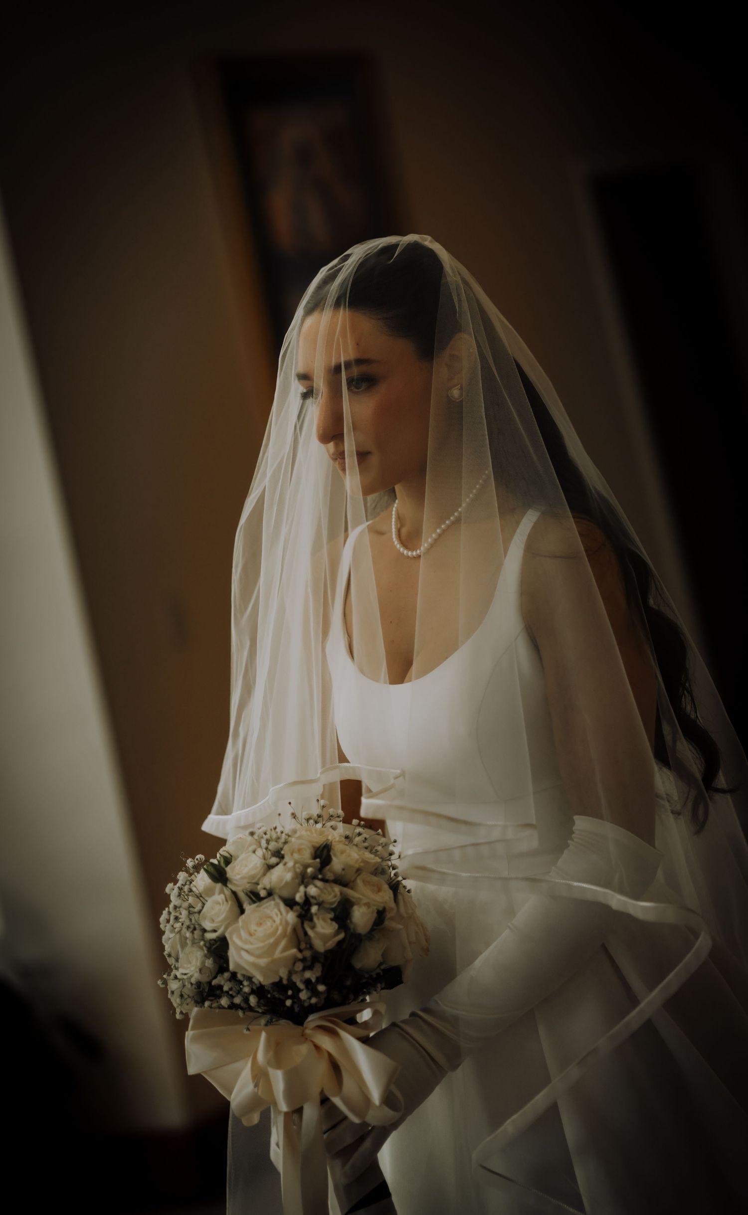 Bride in white wedding dress with flowing veil holds elegant bouquet of white roses in soft indoor lighting.Editorial and Cinematic Wedding