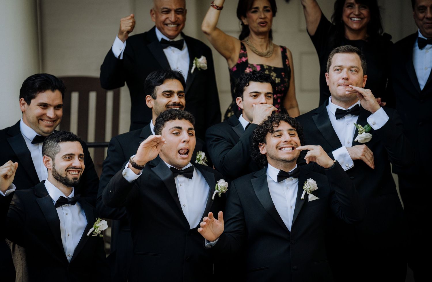 Groomsmen in black tuxedos with boutonnieres strike fun poses on steps at a wedding celebration.