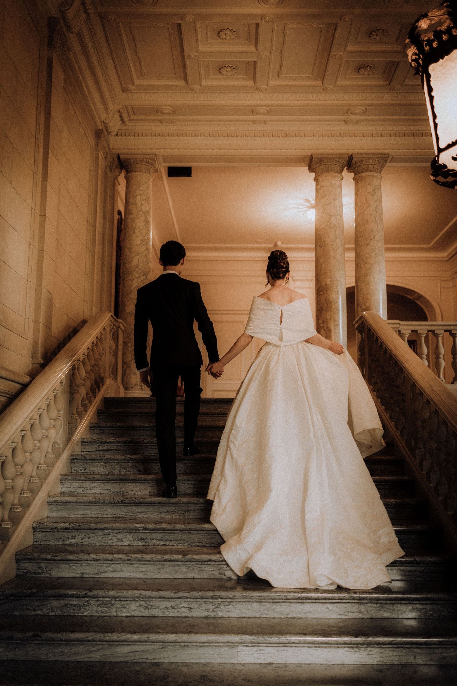 Bride and groom walking up grand marble staircase in elegant venue, bride in flowing white gown holding hands with groom in black suit.