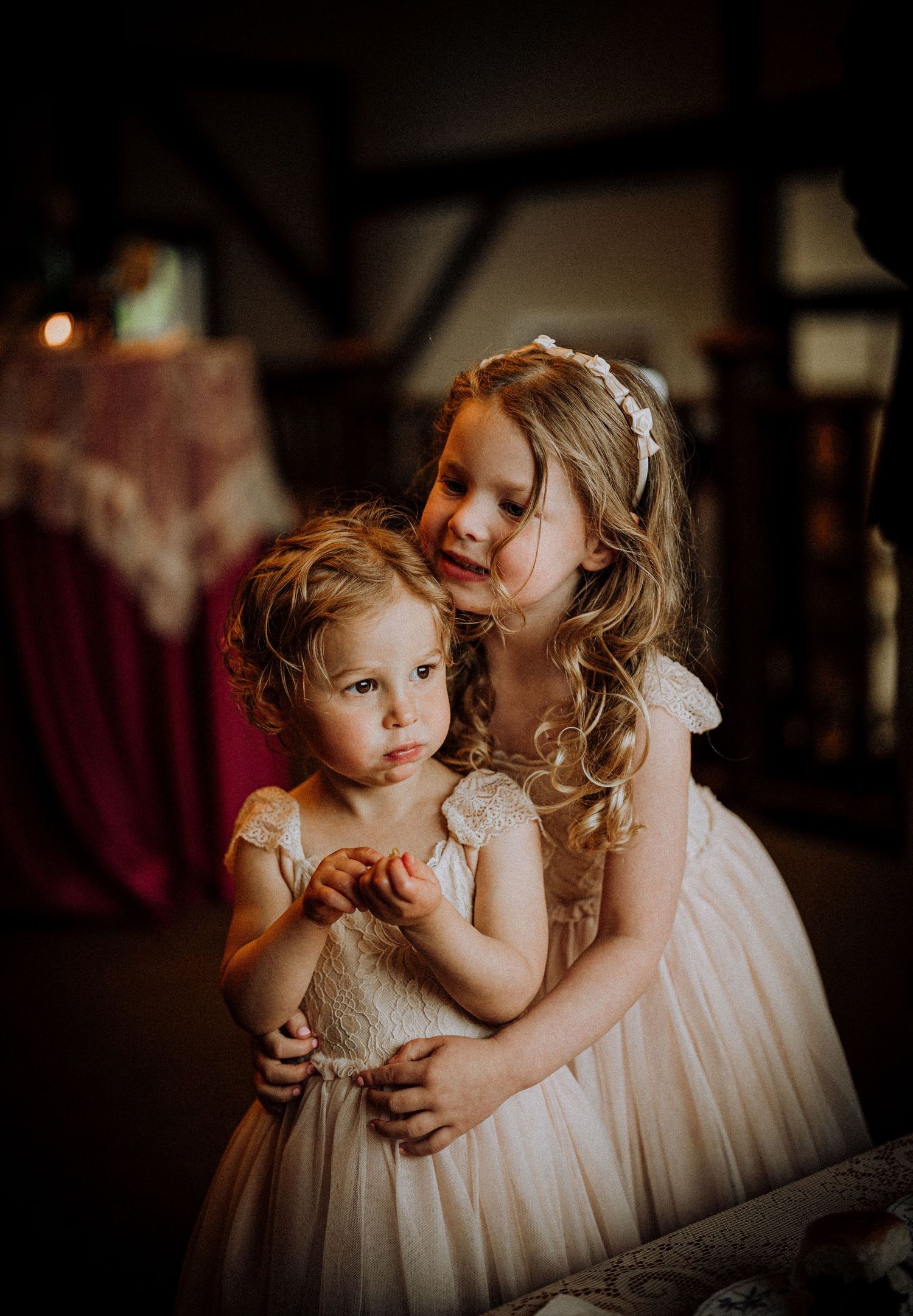 Two young flower girls in white lace dresses embrace tenderly at a wedding reception in a rustic barn venue.