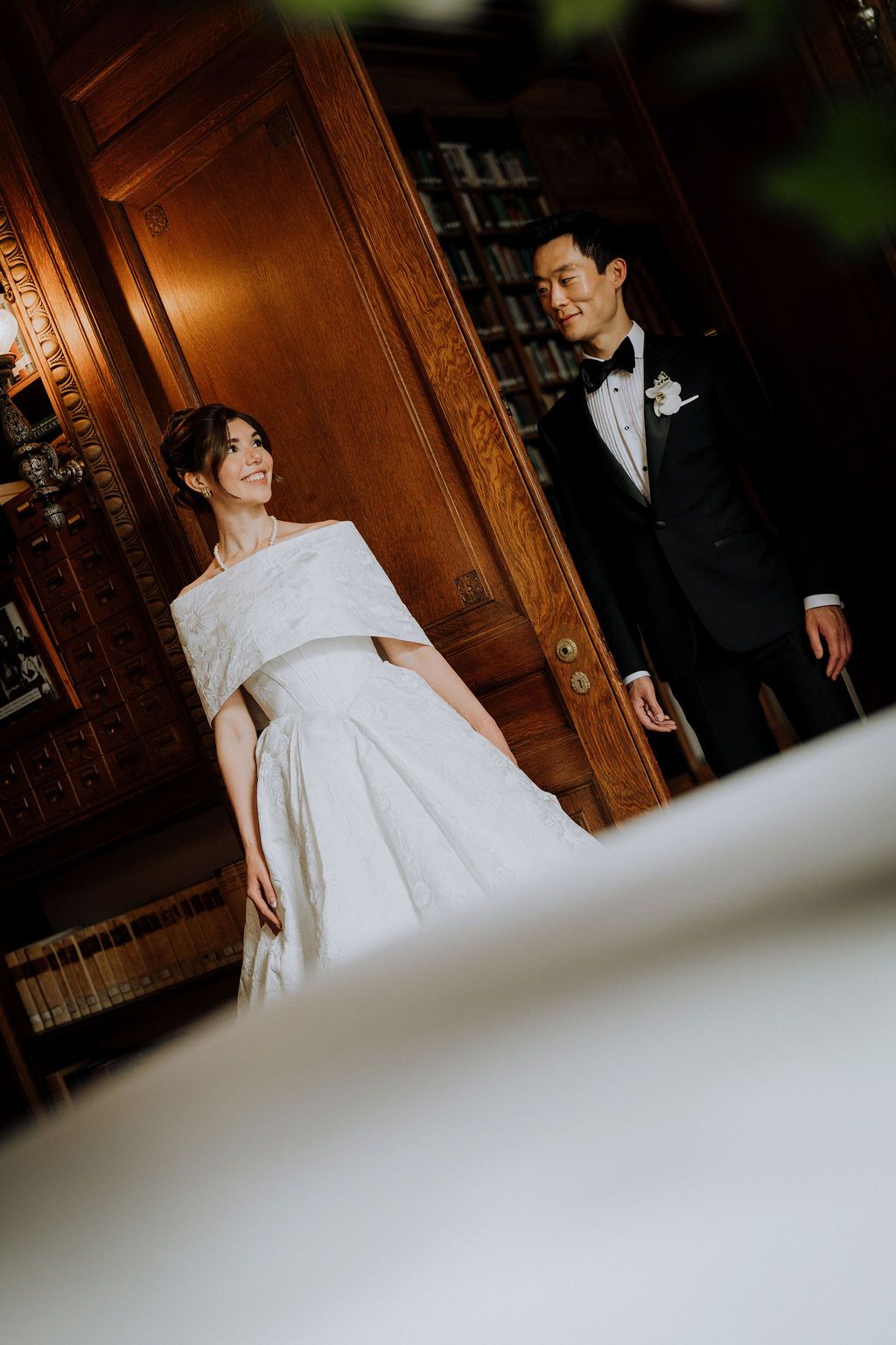 Bride in white off-shoulder gown smiles at groom in black tuxedo in elegant wood-paneled room.