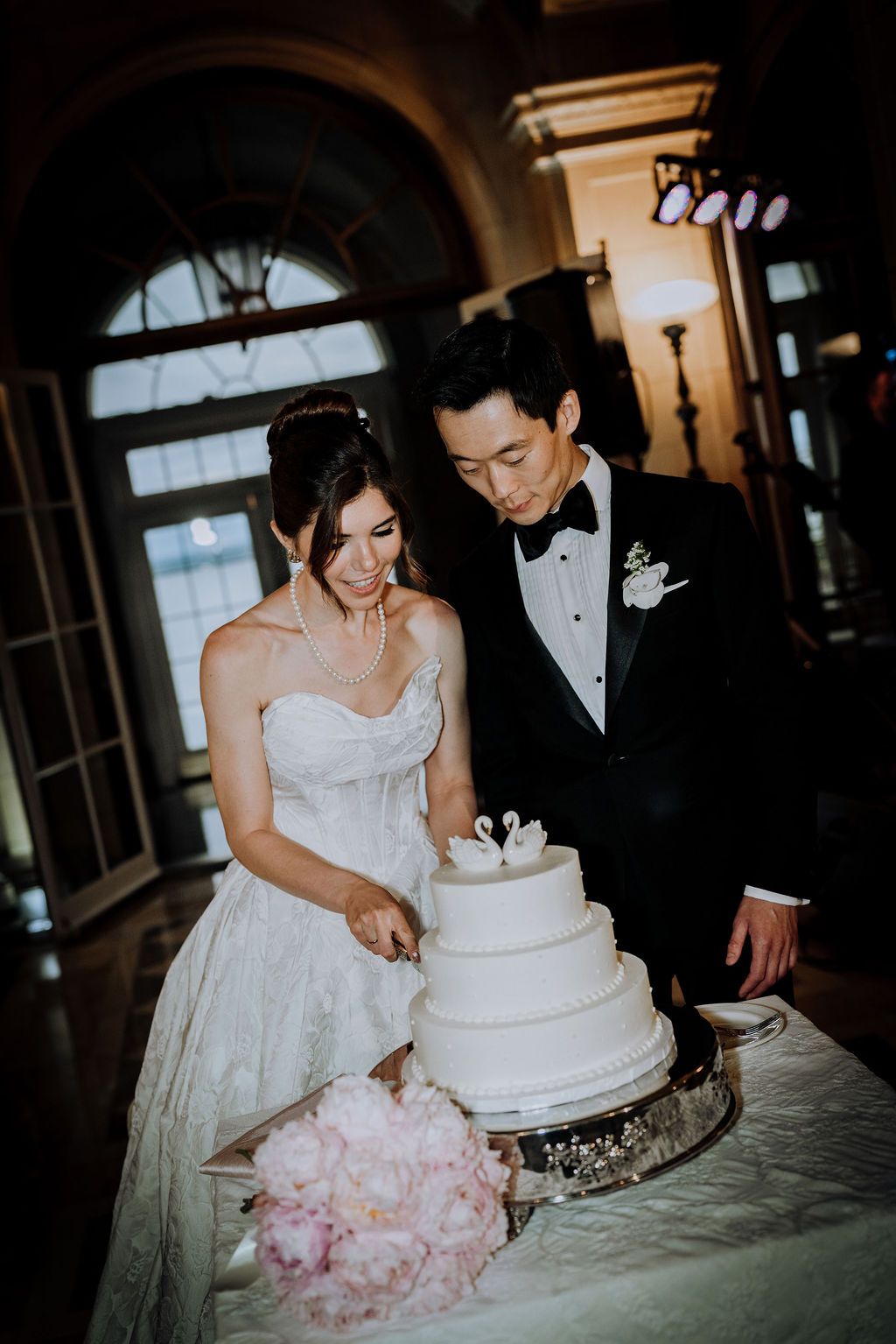 Bride and groom cutting white tiered wedding cake decorated with swan toppers at reception.