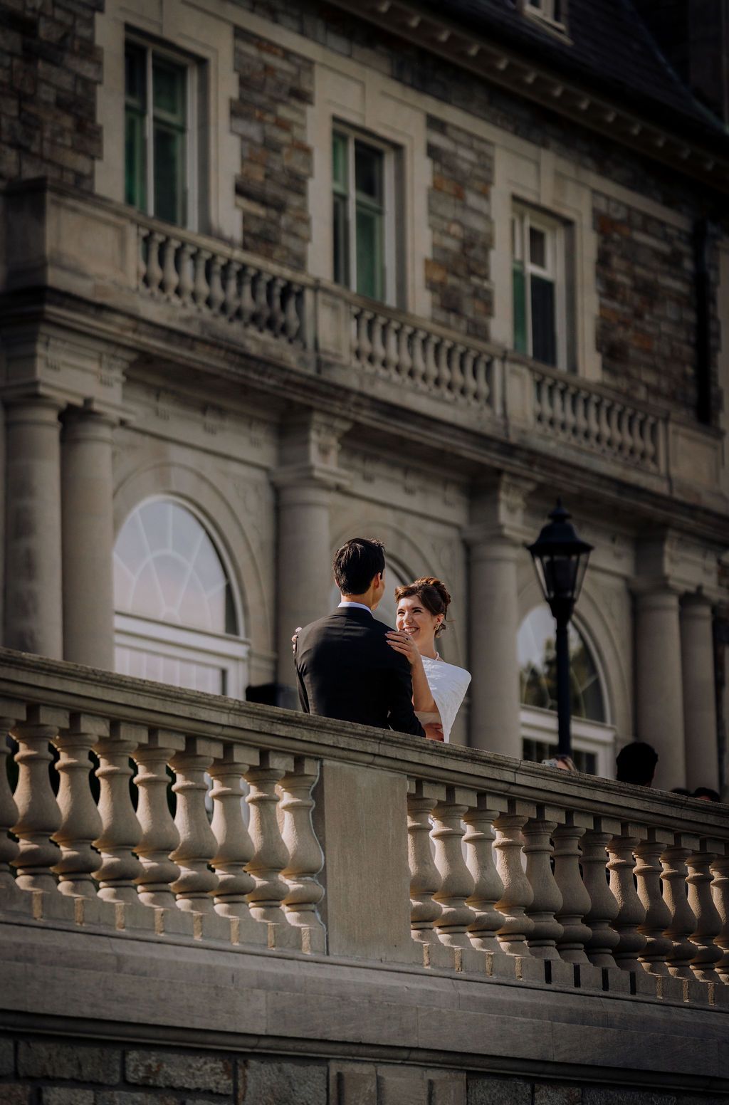 Elegant couple in wedding attire standing on a stone balustrade balcony of a grand historic building.