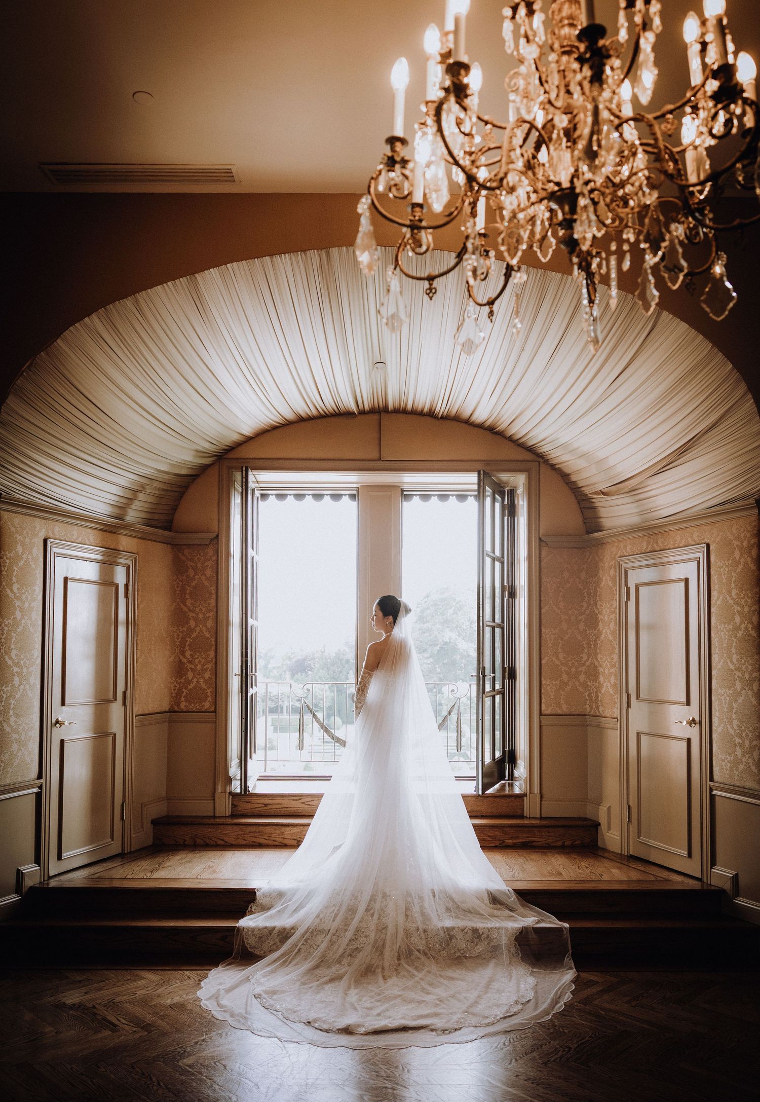 Bride in a long white gown with cathedral veil stands in an elegant arched hallway with crystal chandelier.