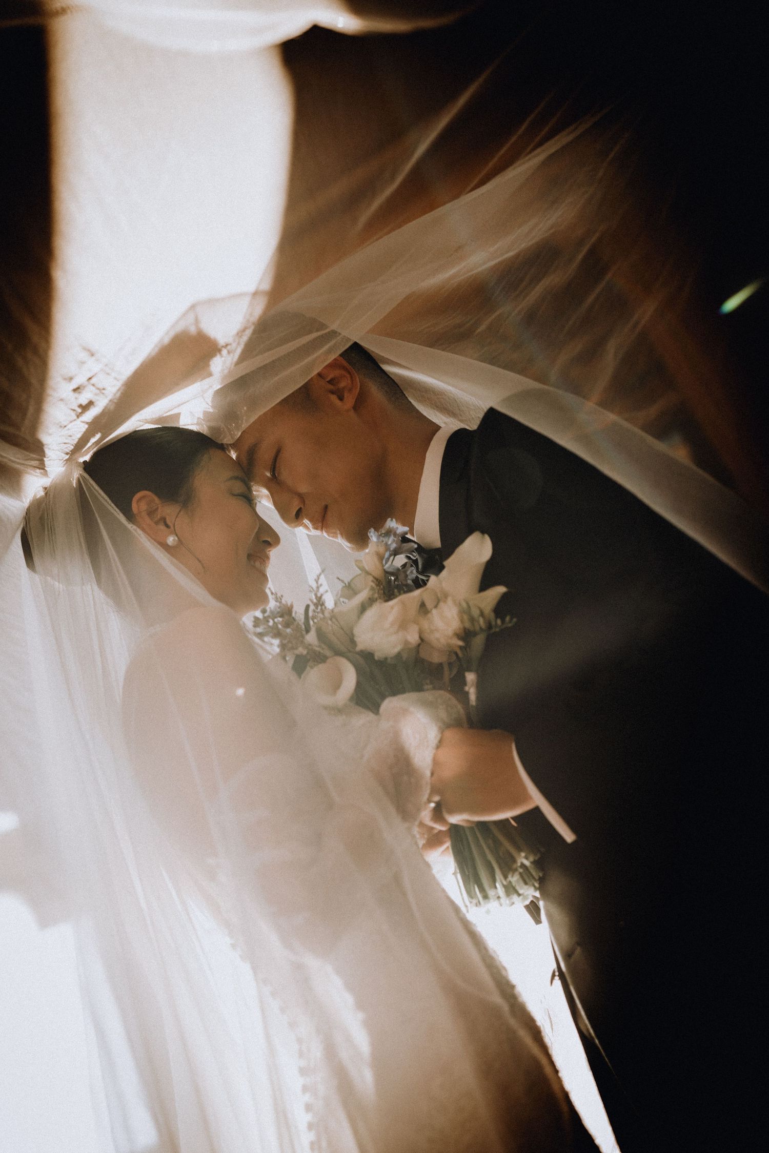 Bride and groom share an intimate moment under a flowing veil, bathed in soft golden light, holding a white floral bouquet.