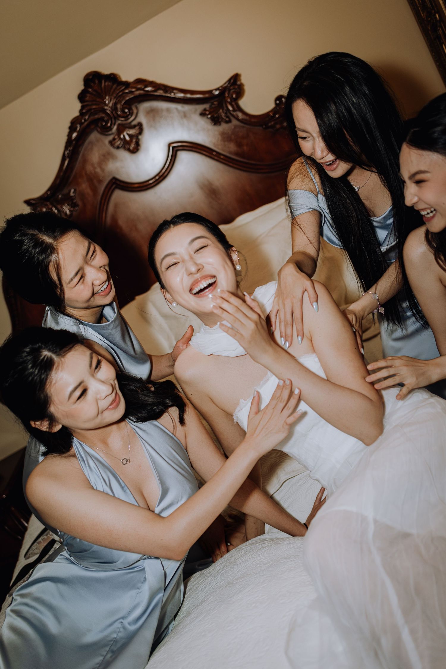 Bride laughing joyfully with bridesmaids in blue satin dresses on a bed during wedding preparations.