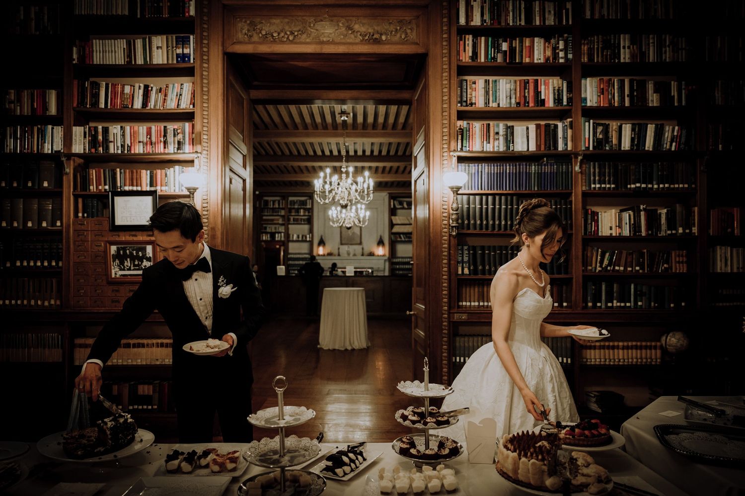 Bride and groom at elegant library wedding reception dessert table surrounded by floor-to-ceiling bookshelves and chandelier.