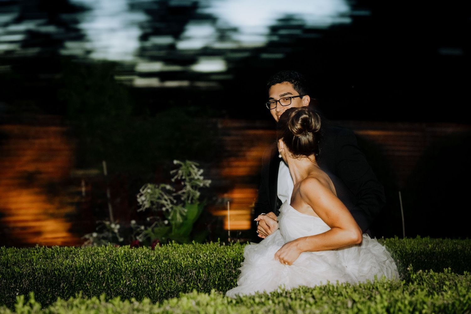 A bride and groom share a romantic embrace on green grass at night, with warm garden lighting behind them.