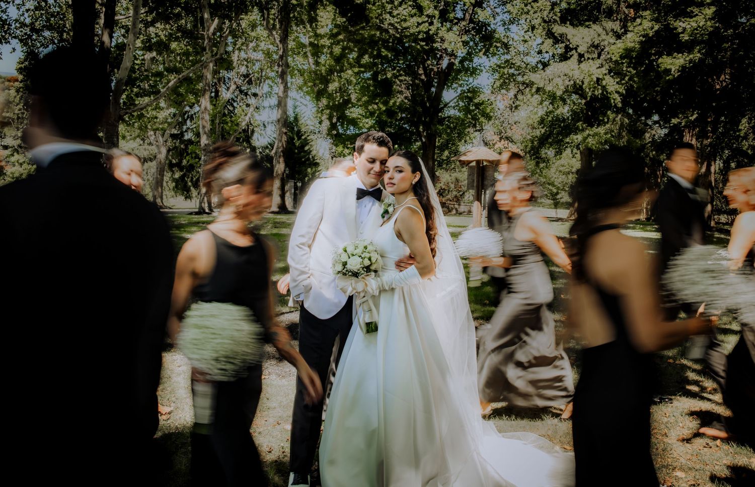 Bride and groom pose outdoors surrounded by blurred wedding party guests in a sunlit forest setting.Editorial and Cinematic Wedding photos