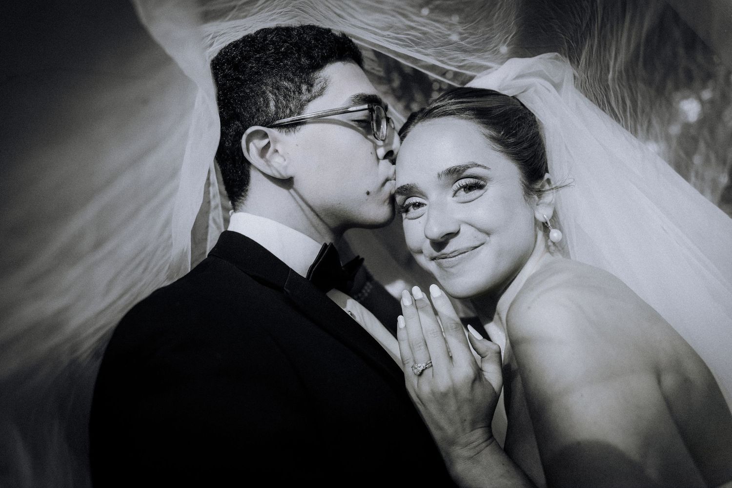Black and white wedding photo of groom kissing bride on cheek as she smiles, draped in a flowing veil.