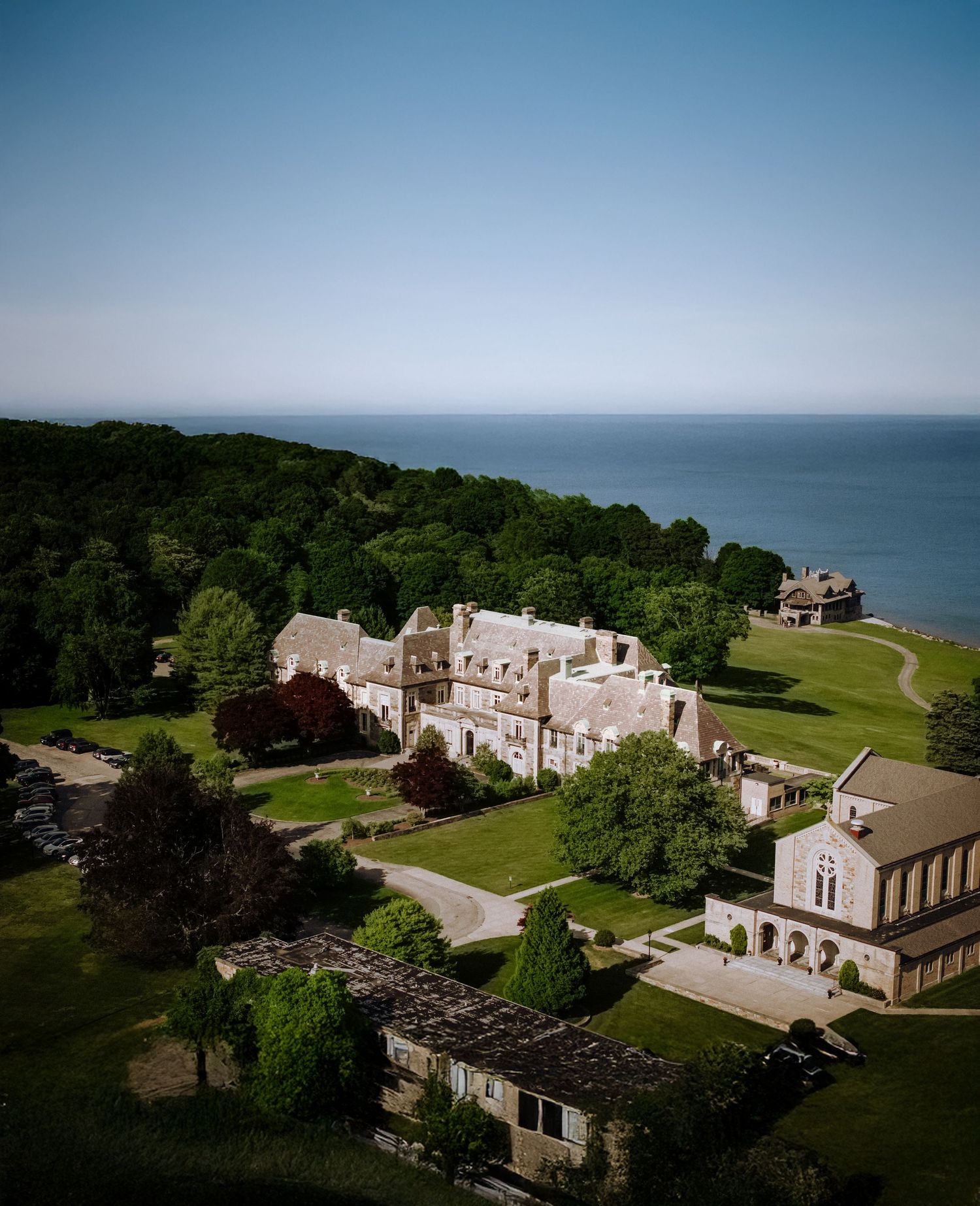 Aerial view of a grand historic estate surrounded by lush trees with scenic ocean views under a clear blue sky.