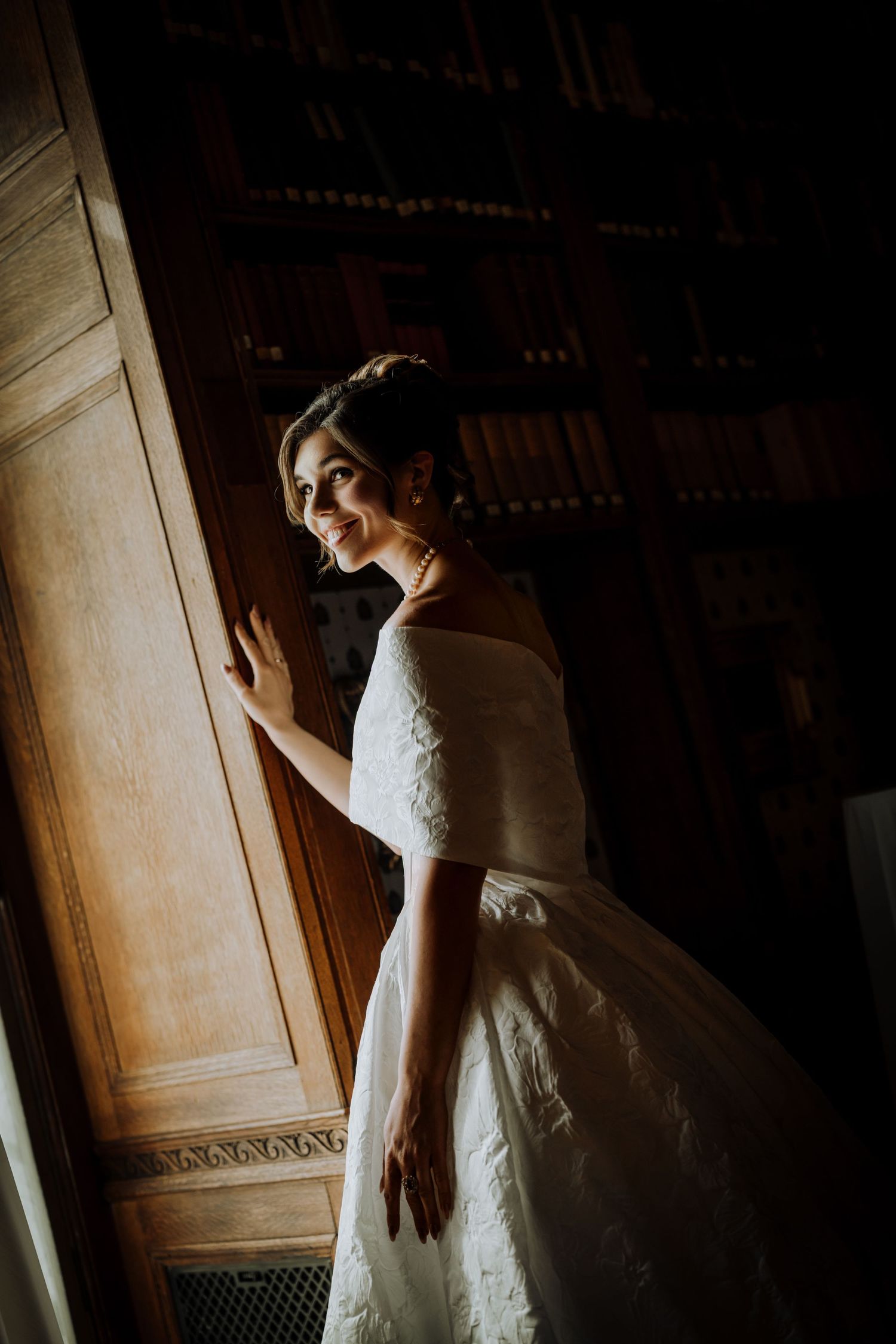 Bride in white off-shoulder lace gown smiling beside wooden door in dimly lit elegant interior.