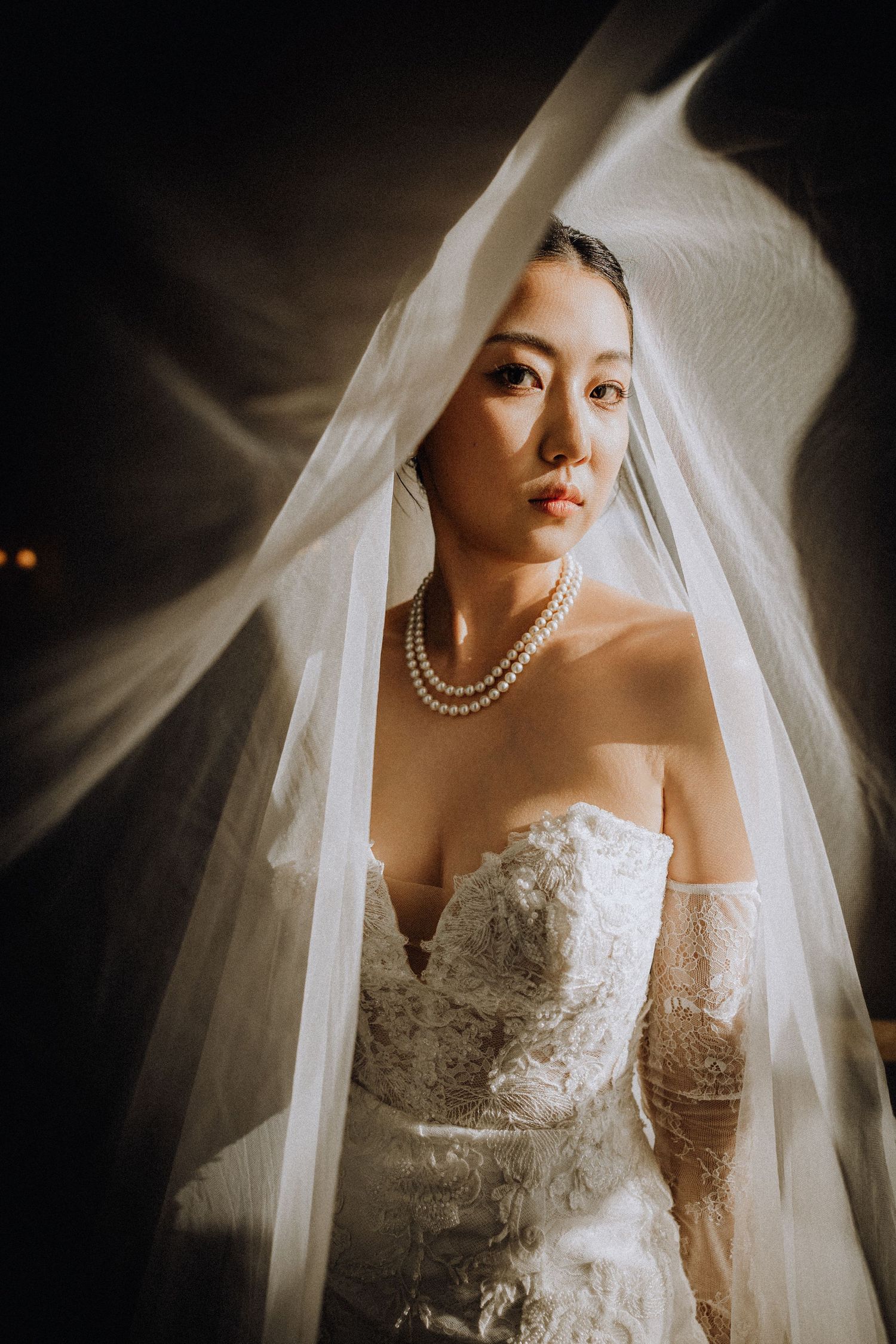 Bride in lace off-shoulder gown and pearl necklace draped in flowing white veil, elegant bridal portrait.
