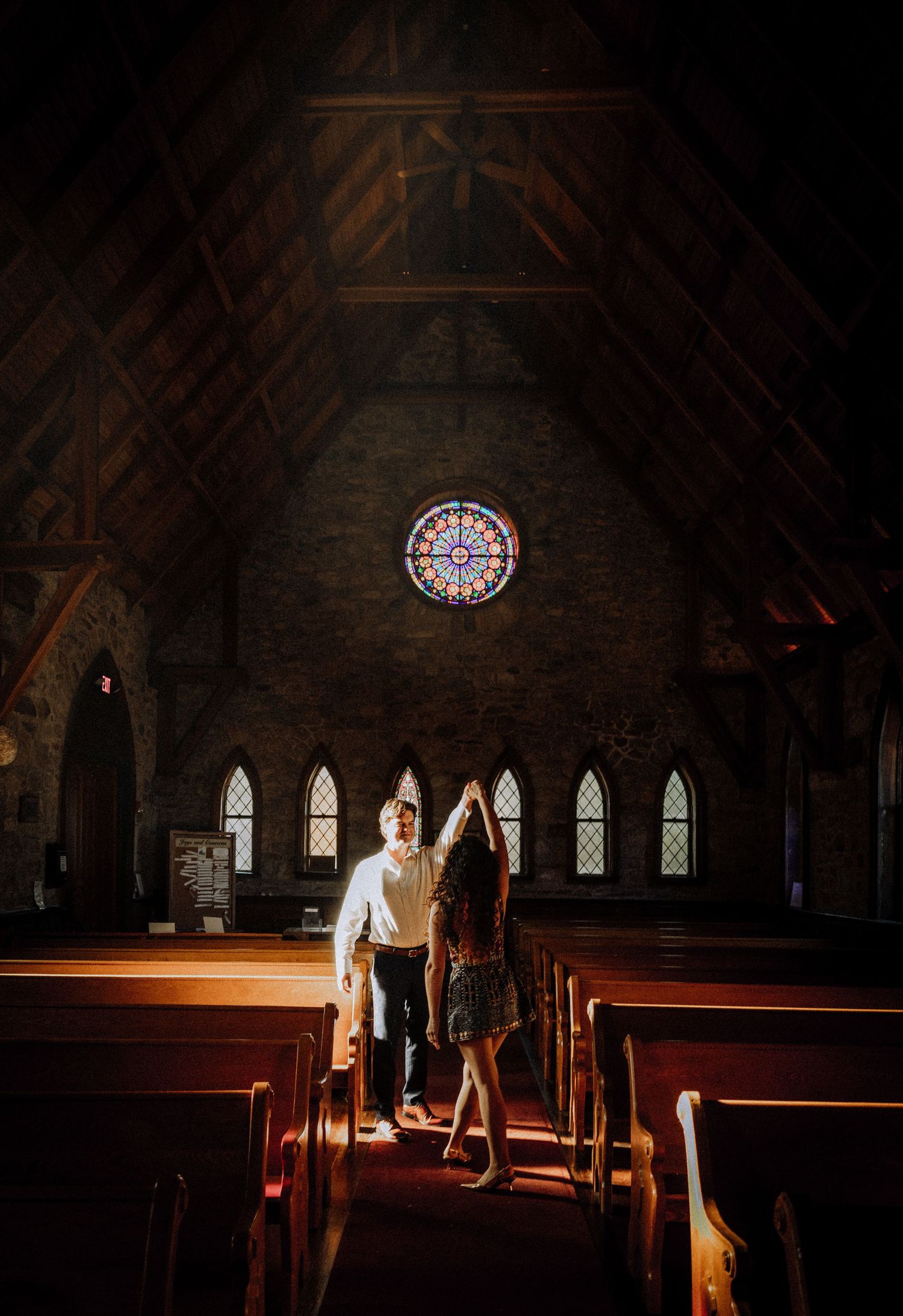 A couple standing in church aisle beneath vaulted wooden ceiling and stained glass.