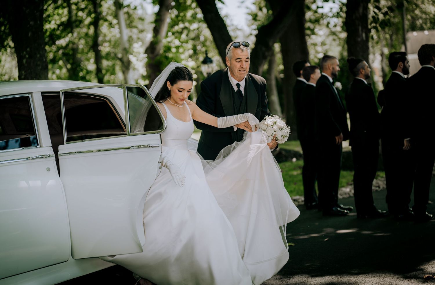 Wedding party gathered outdoors in elegant attire with white limousine in background.