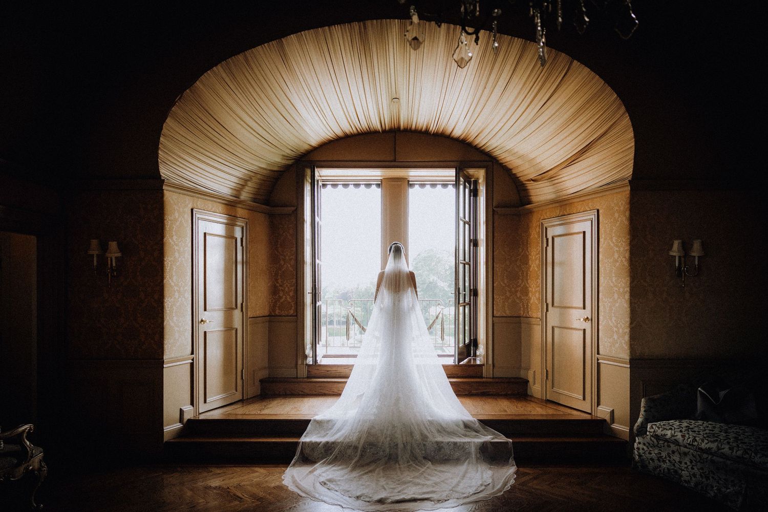 Bride in elegant white gown with long veil stands backlit in arched doorway of rustic venue.