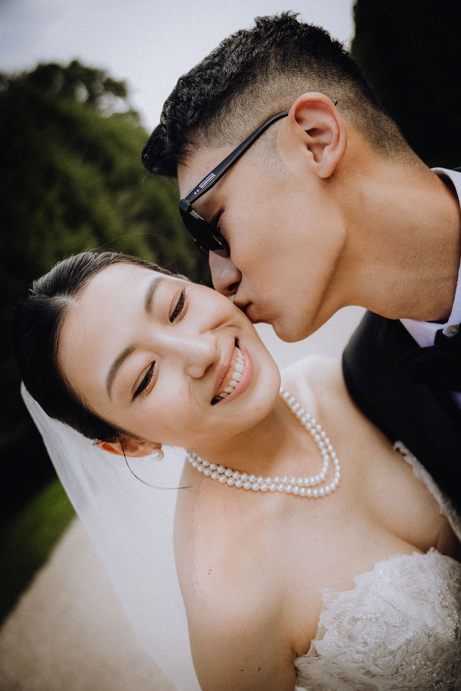 Groom in black suit kisses smiling Asian bride wearing pearl necklace and white veil outdoors.