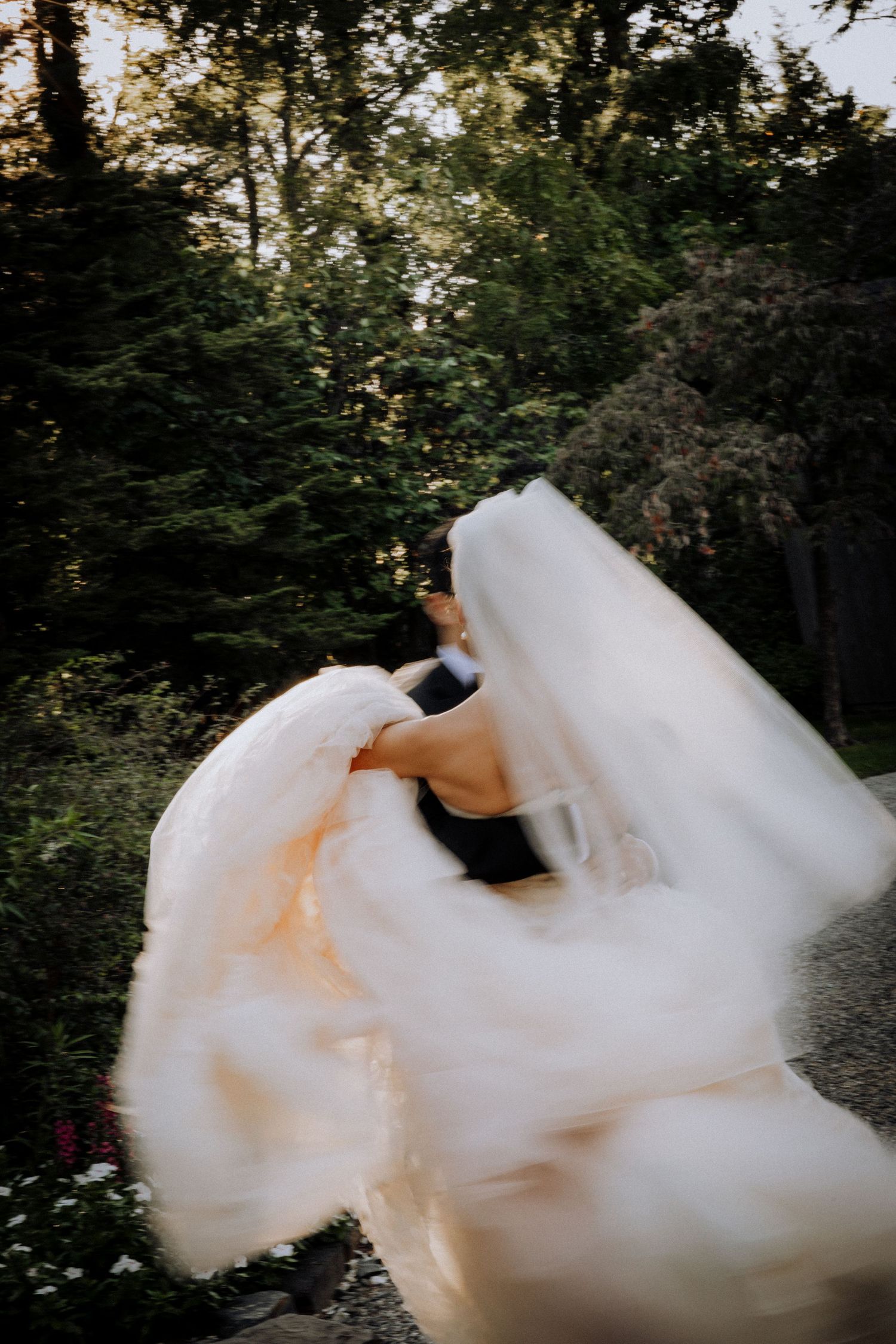 Bride twirling in flowing white wedding dress and veil outdoors surrounded by lush green trees.