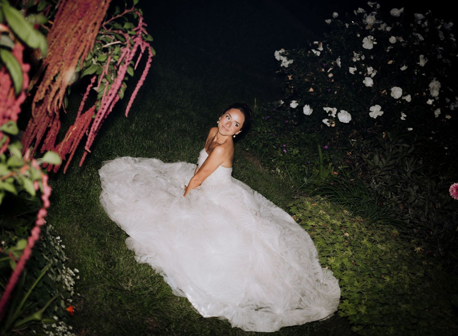 Bride in a white ball gown sitting on green grass at night surrounded by pink and white floral decorations.