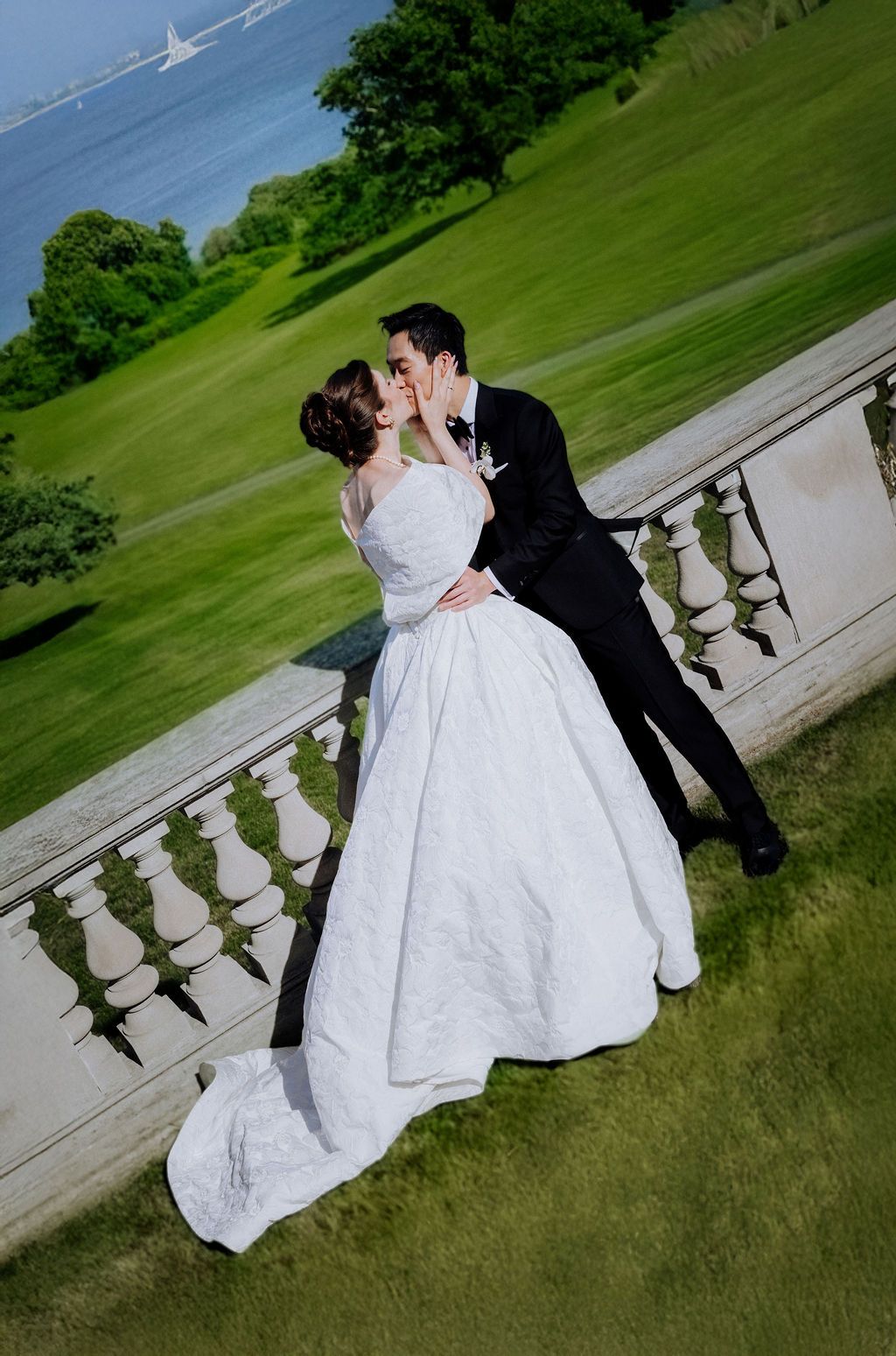 Bride in a ballgown and groom in a tuxedo share a kiss on a stone balustrade overlooking green lawns.