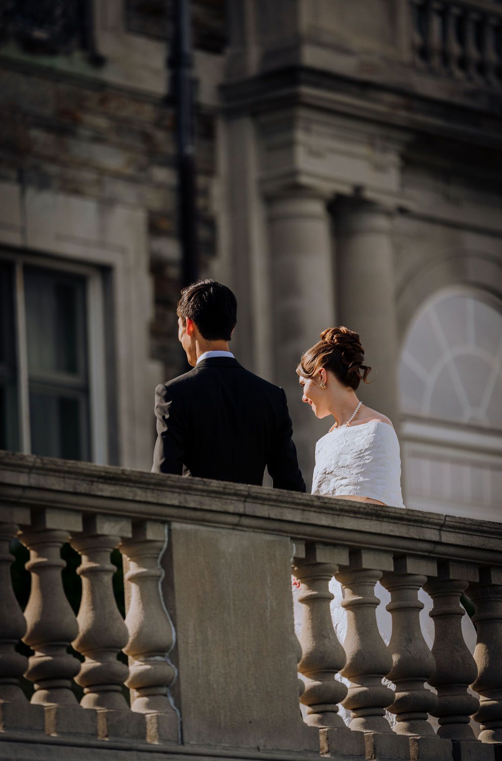 A bride and groom stand together on an ornate stone balcony, viewed from behind, in elegant wedding attire.