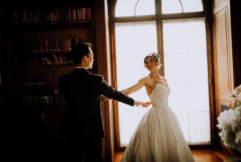 Bride and groom share a romantic first dance near a sunlit window in an elegant library setting.