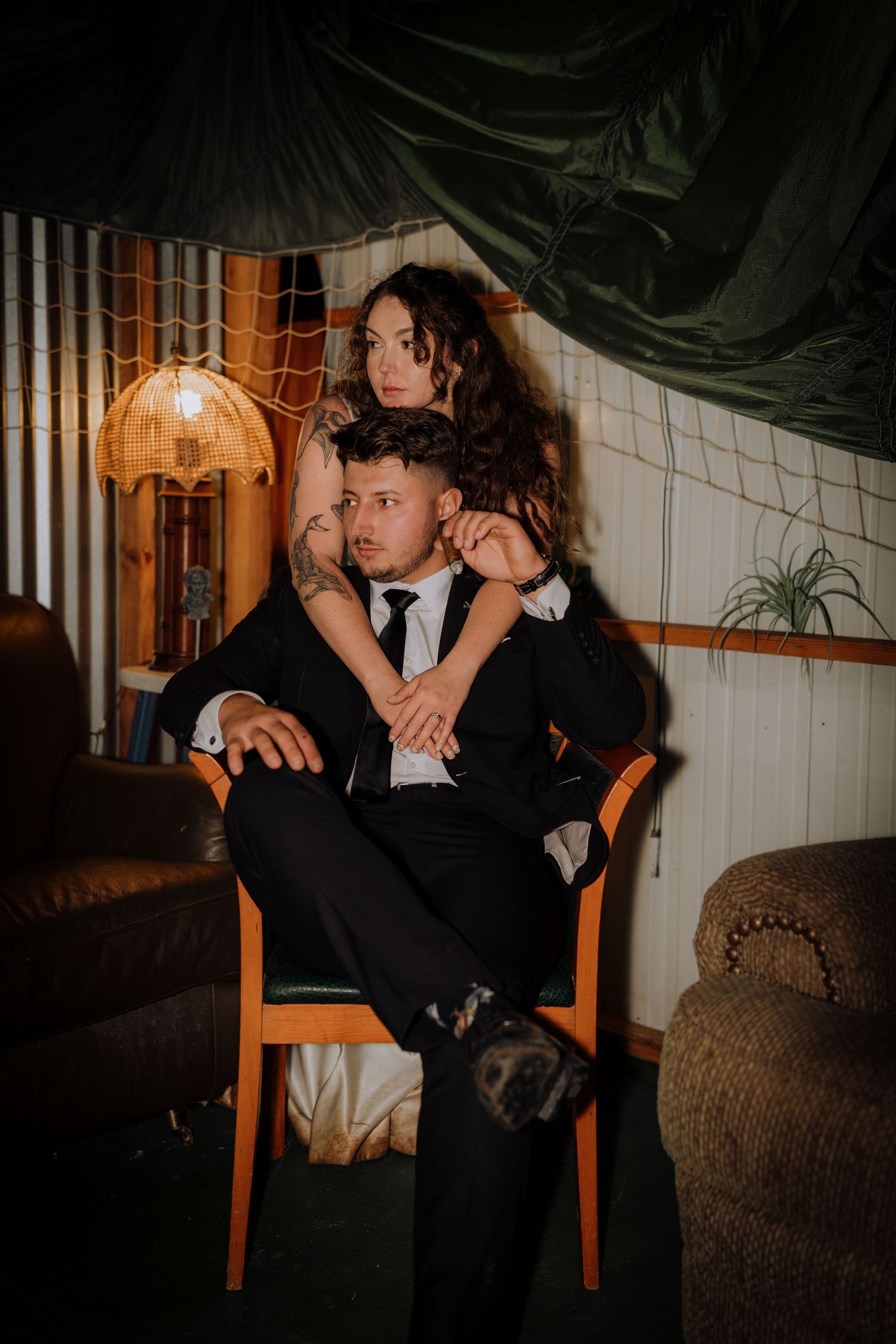 Couple sharing intimate moments on wooden chairs in a dimly lit setting.