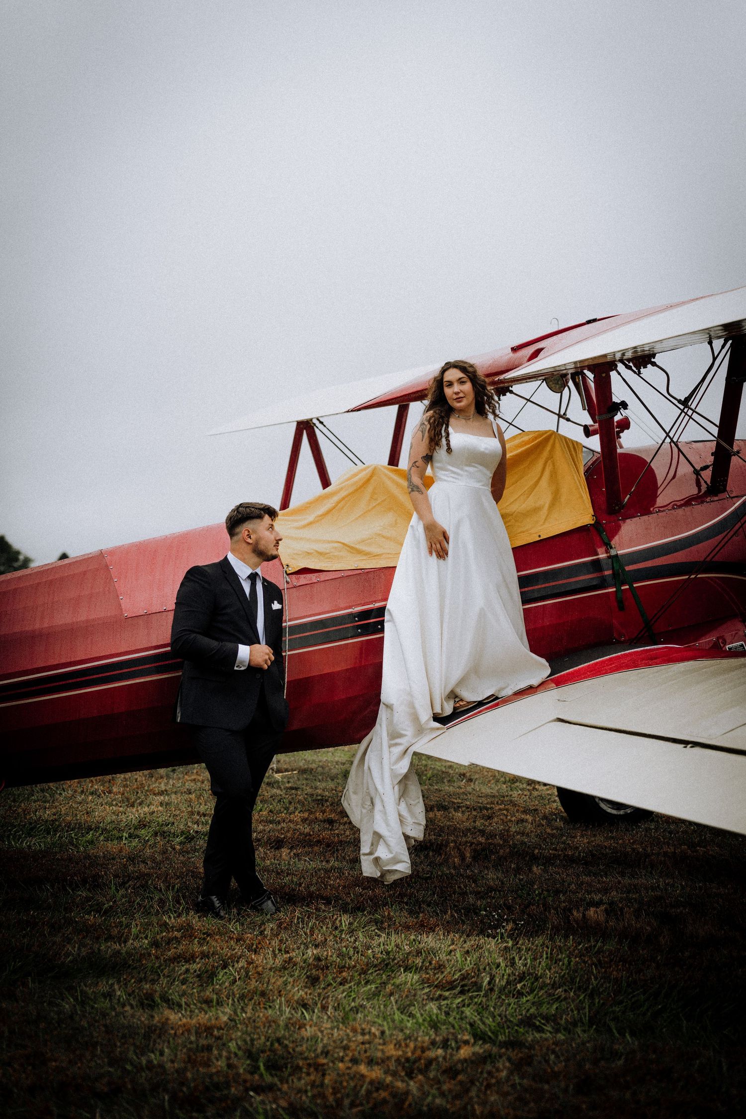A wedding couple poses dramatically with a vintage red biplane in an open field.