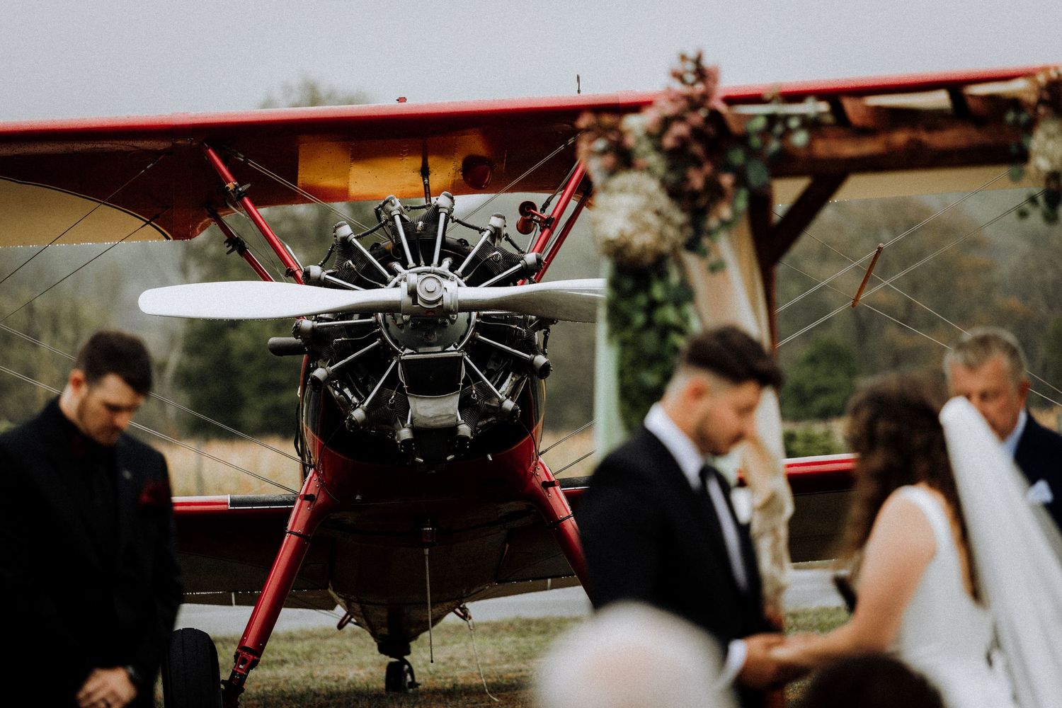 A vintage red aircraft with prominent propeller serves as backdrop for wedding party photos.