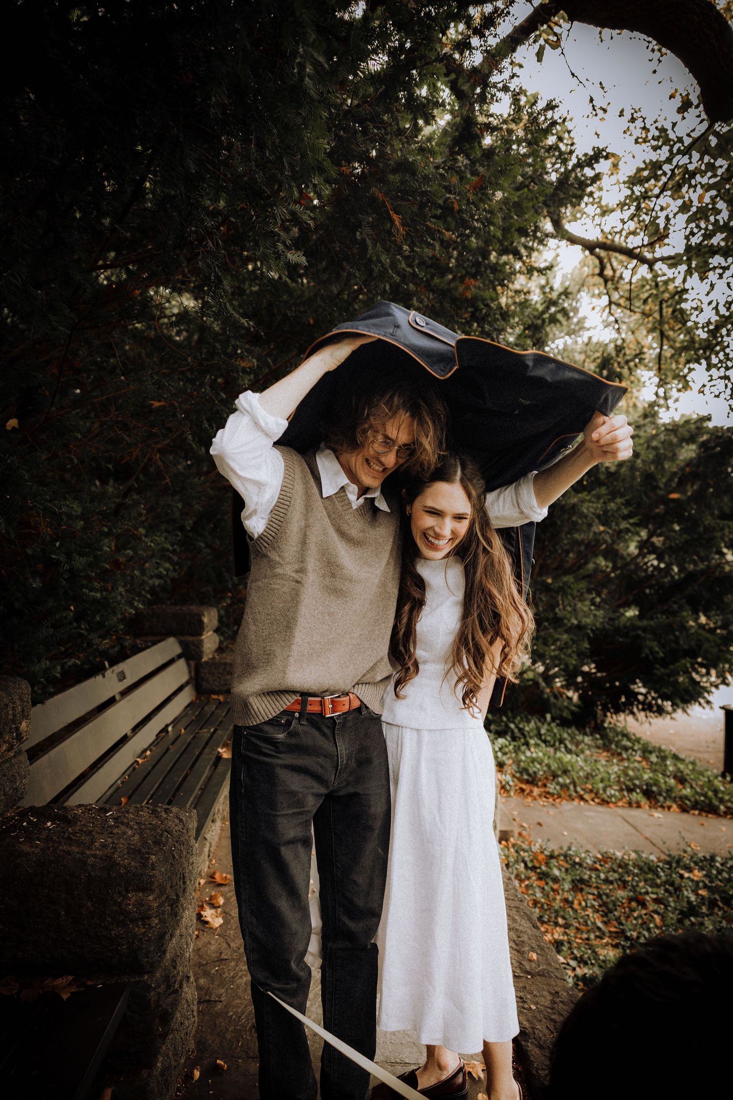 A couple dressed in white and brown embraces outdoors against a natural backdrop with trees.