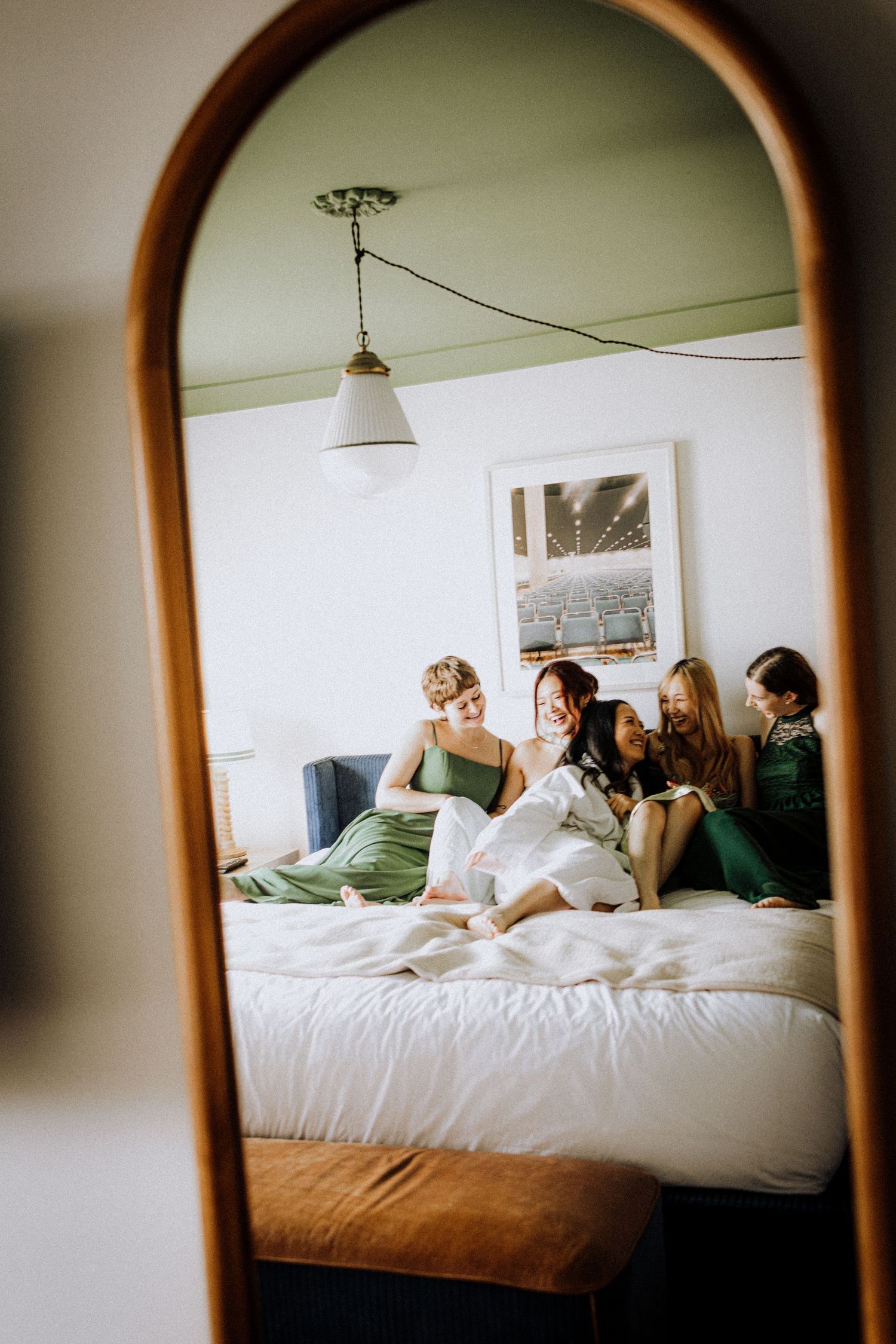 Four bridesmaids in green dresses sit together on a white bed, reflected in an arched wooden mirror.