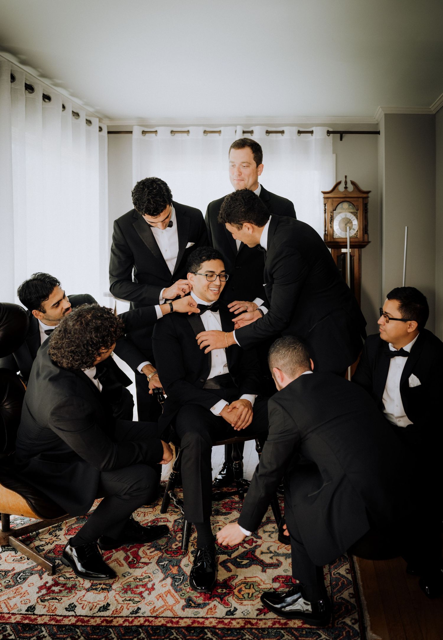 Groomsmen in black tuxedos helping groom get dressed before wedding ceremony in elegant room.